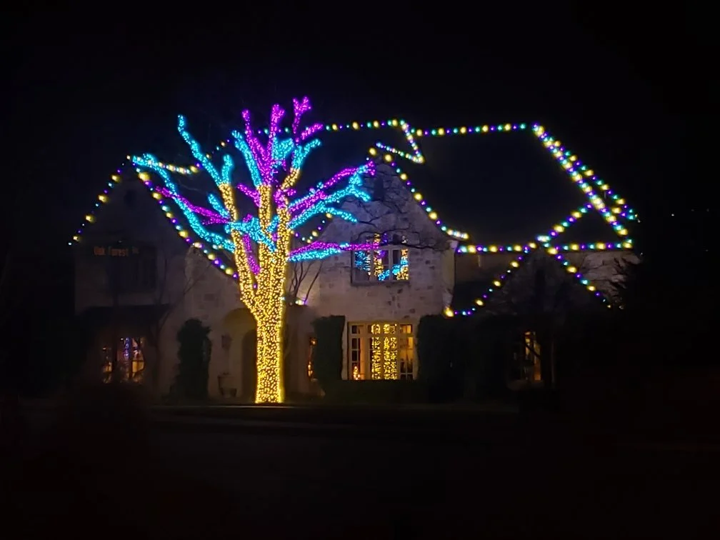 A house decorated with colorful holiday lights, including a large tree wrapped in yellow lights and adorned with blue and purple lights on the branches, with additional string lights creating a large bow-shaped outline in the sky.