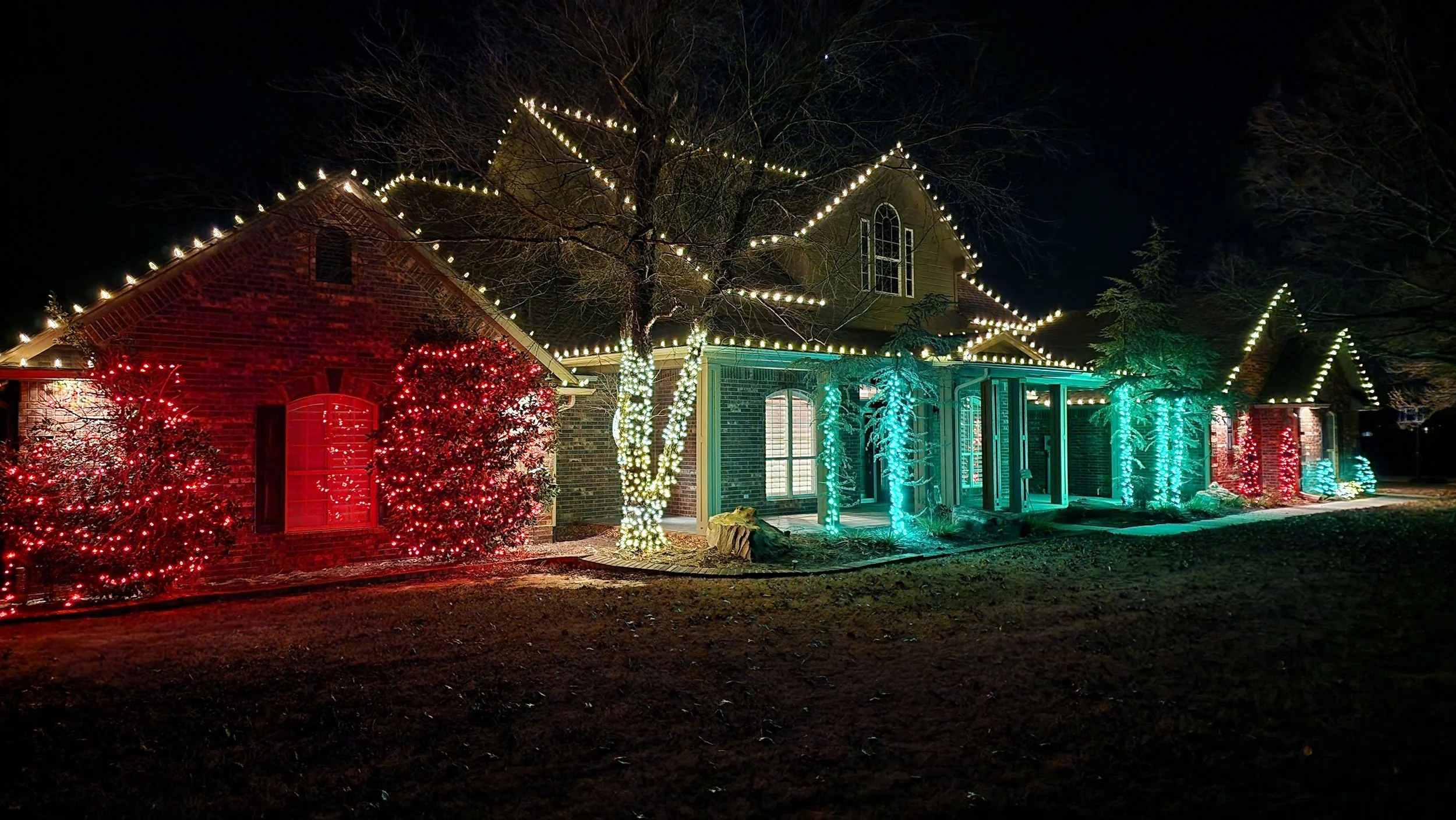 A house decorated with Christmas lights at night, with red, green, and white lights outlining the roof and wrapped around trees, creating a festive holiday display.