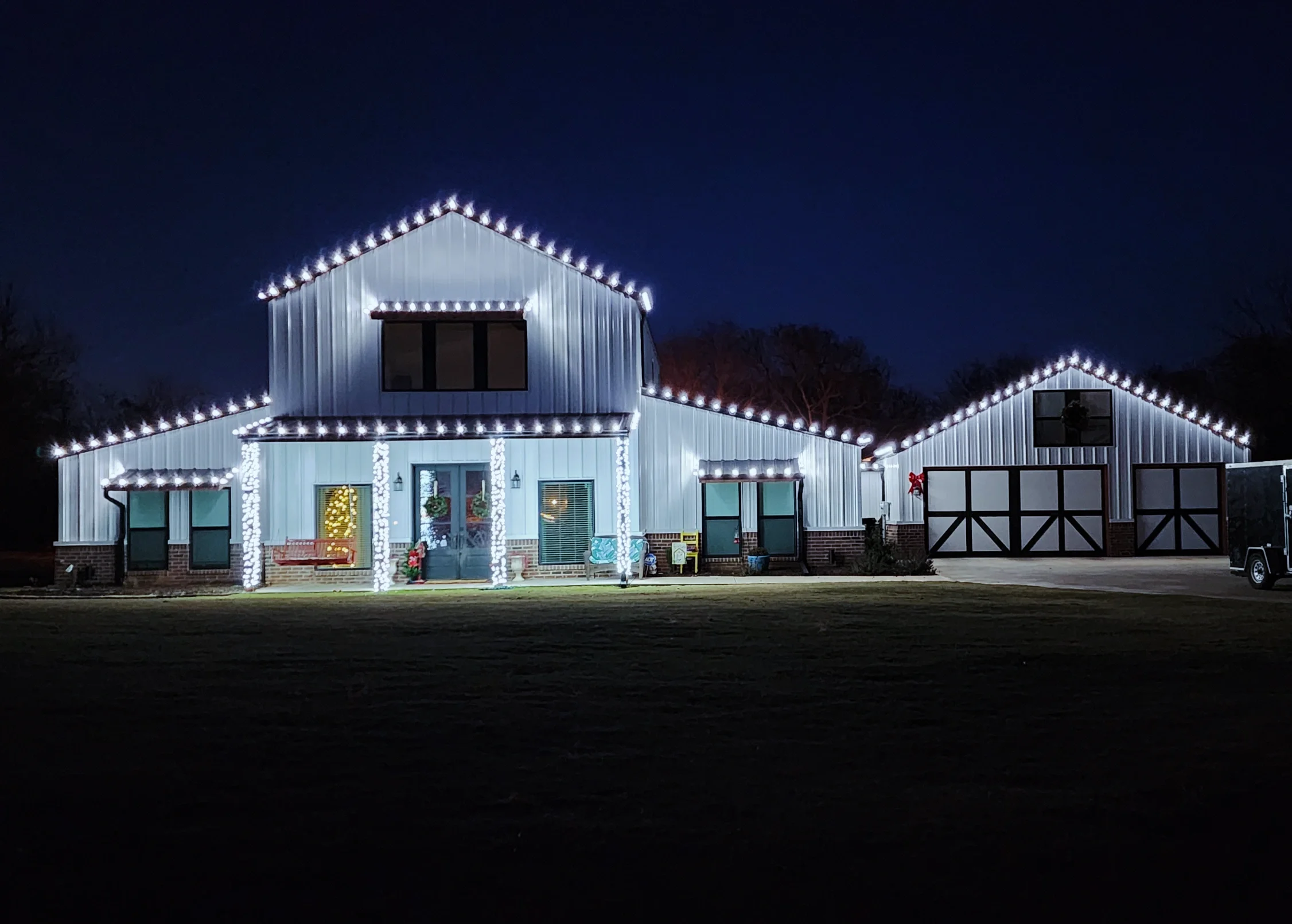 A house decorated with white Christmas lights, illuminated at night, with a two-story main building and attached garage or guest house, and holiday wreaths and decorations visible through the windows.