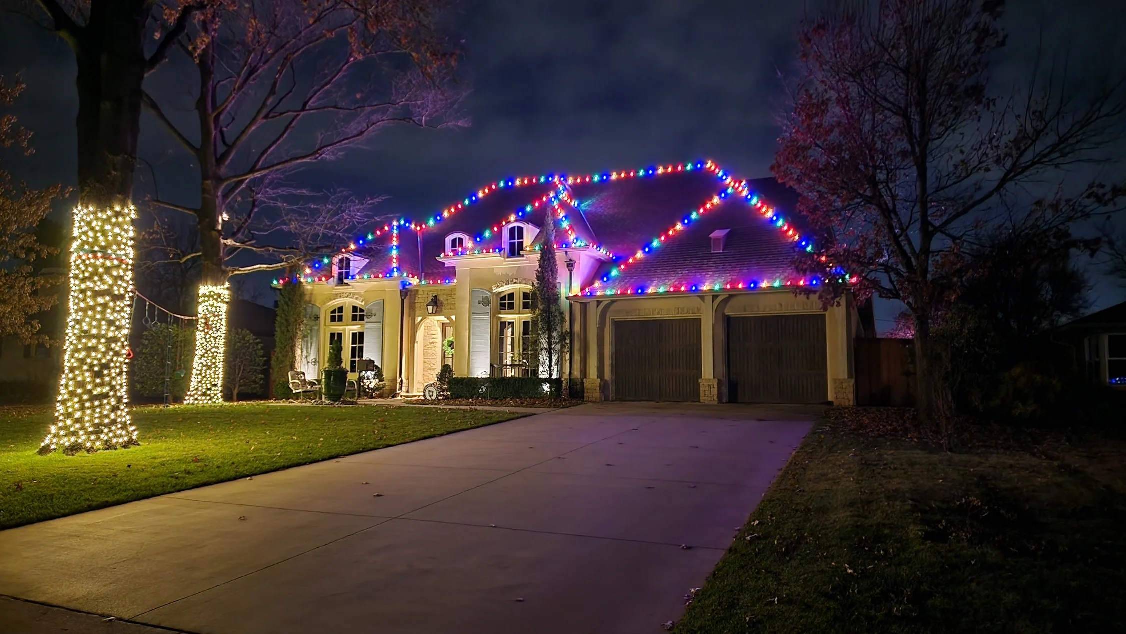 House decorated with multicolored Christmas lights on the roof and white lights wrapped around trees, nighttime scene with a driveway and manicured lawn.