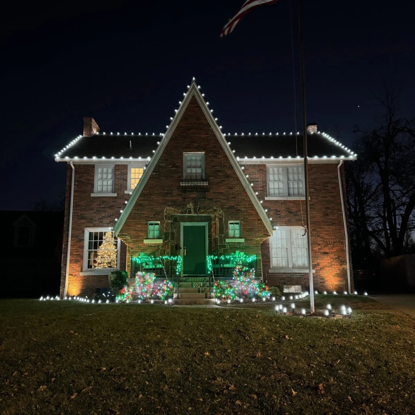 A brick house decorated with Christmas lights at night, including a small decorated Christmas tree in the window, illuminated bushes on the front porch, and an American flag flying in the yard.