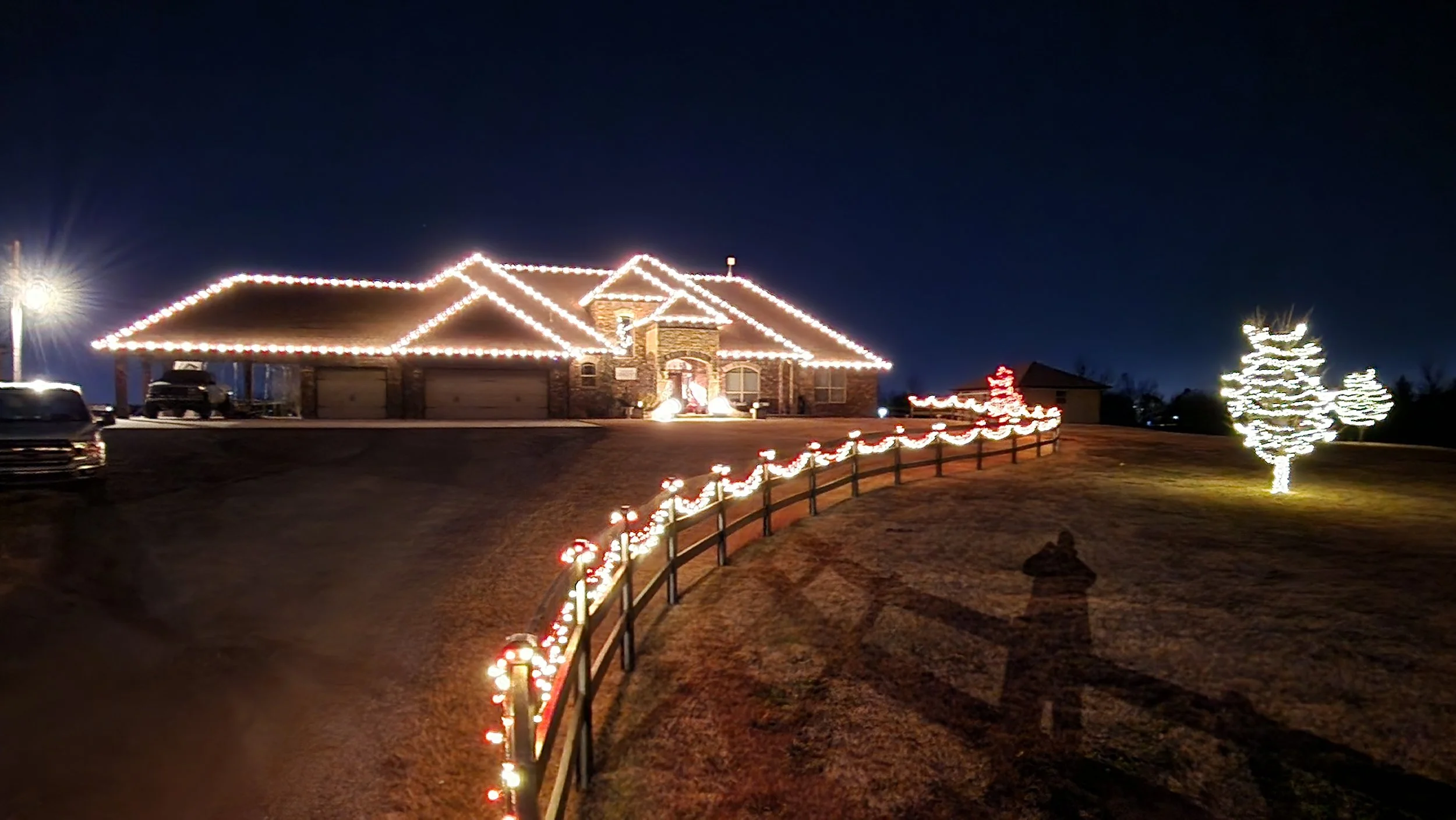 House decorated with Christmas lights, with fencing outlined in lights and trees decorated with white lights, during the night.