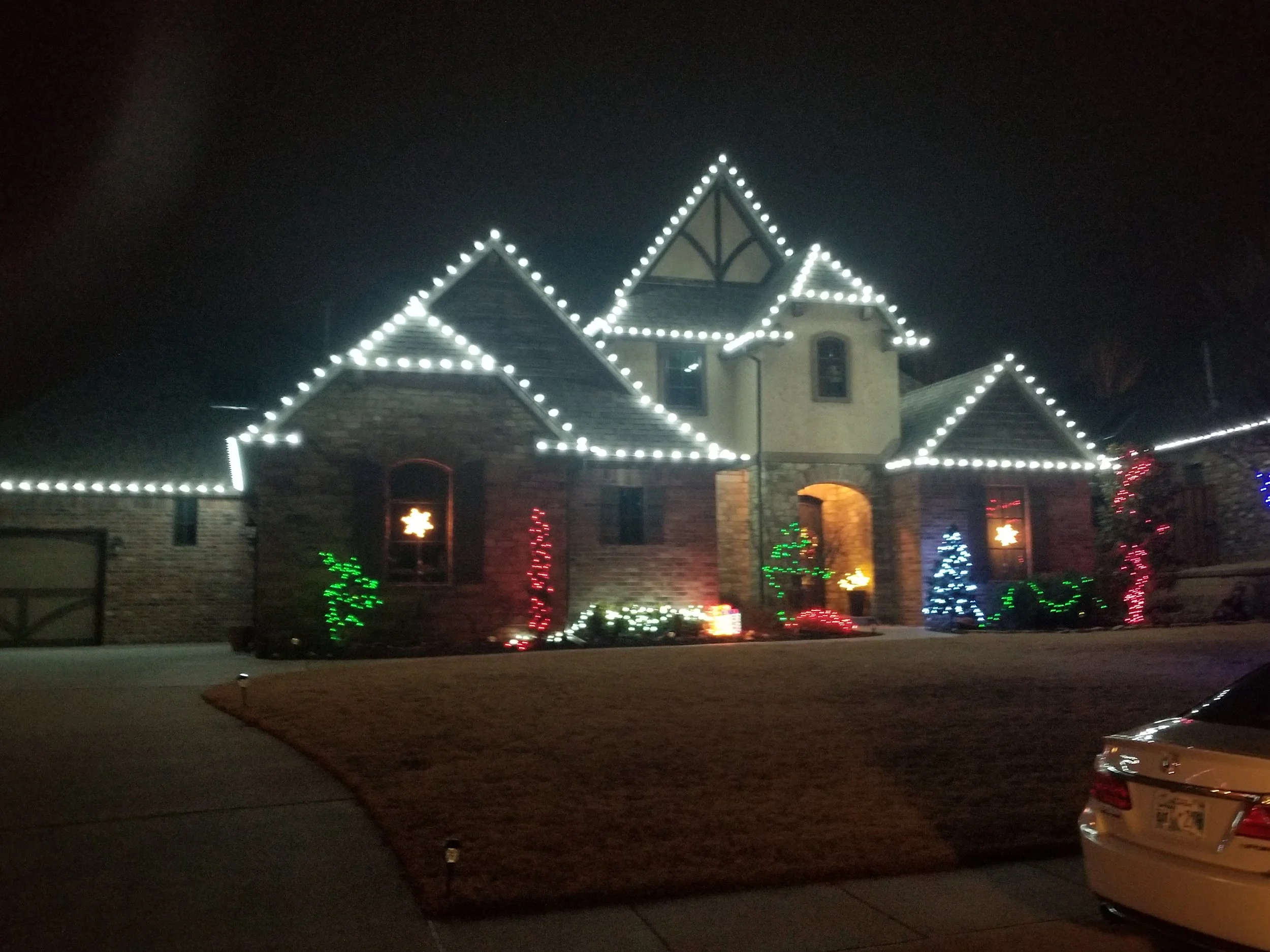 A house decorated with Christmas lights at night. The house has white lights outlining the roof and windows, with colorful lights on the bushes and trees in the yard.