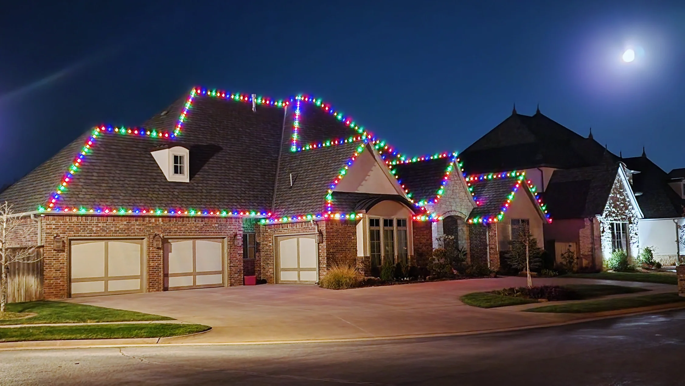 A large brick house decorated with colorful Christmas lights along the roofline, with a full moon in the clear night sky.