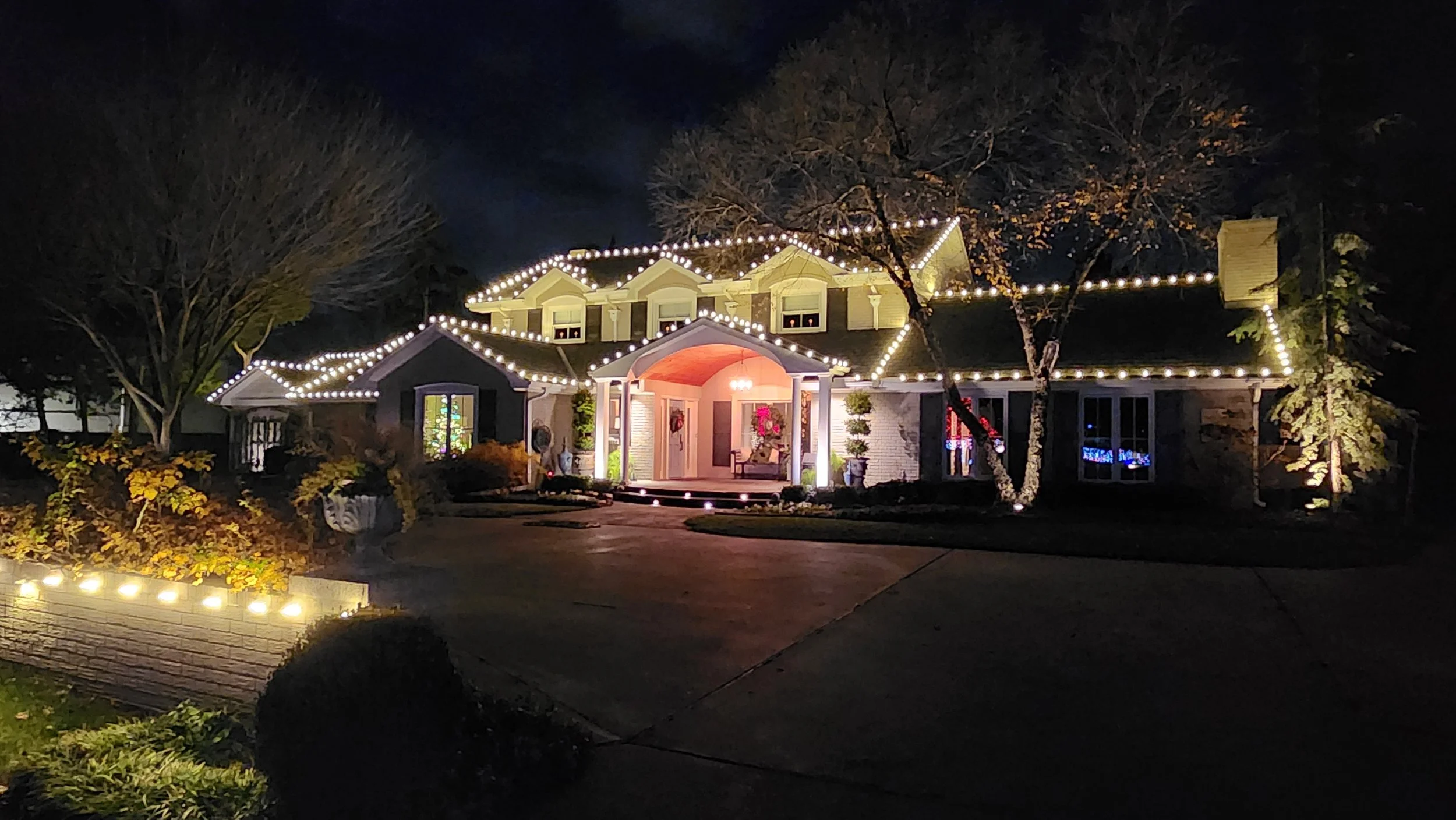 A house decorated with holiday Christmas lights at night, featuring strung lights on the roof and a wreath on the front door.