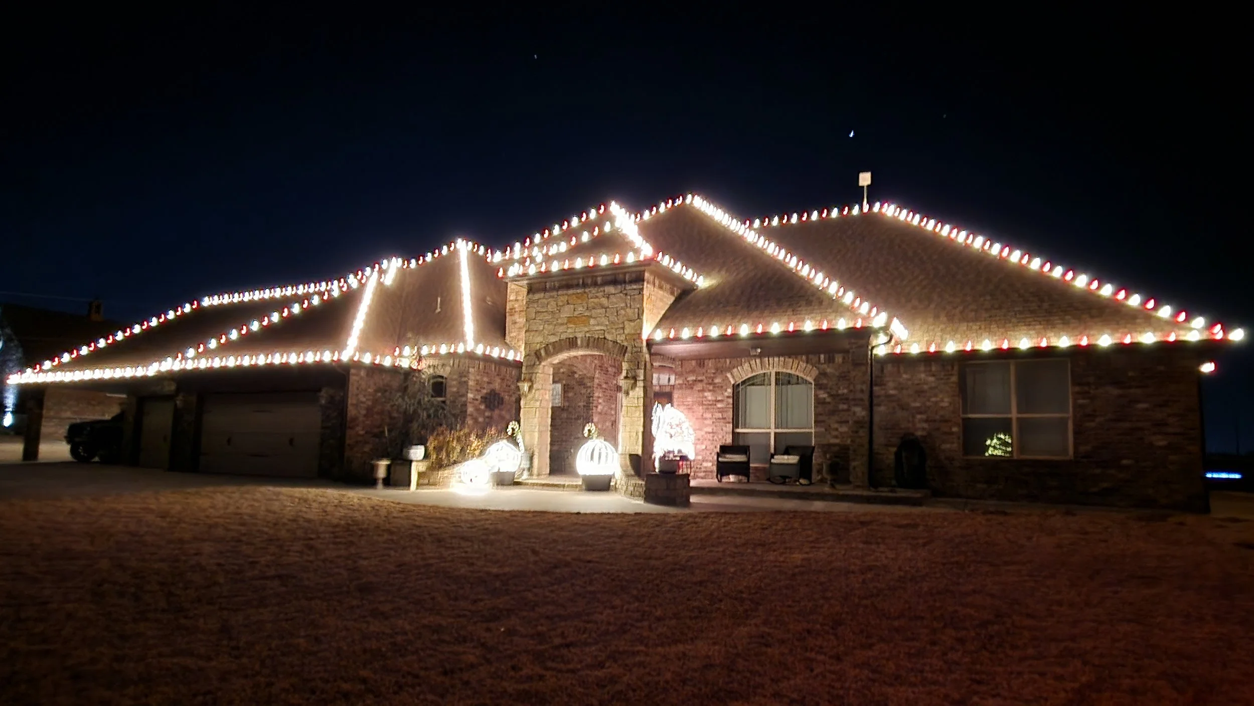 A house decorated with Christmas lights outlining the roof and roof edges at night. There are illuminated decorative pumpkins near the entrance, and the house has a brick exterior with a stone chimney. The yard is dark and the sky is clear with a few