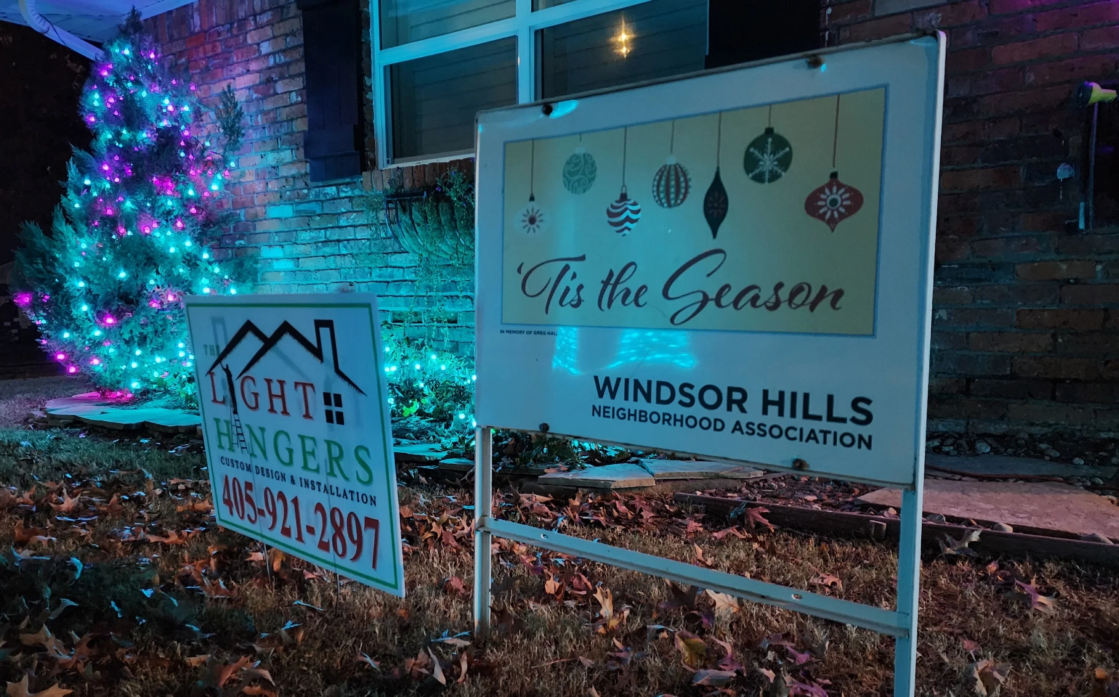 Decorated Christmas tree with multicolored lights next to two signs in a yard at night, one reads '’Tis the Season' with hanging ornament graphics, and the other advertises light hangers with a phone number.