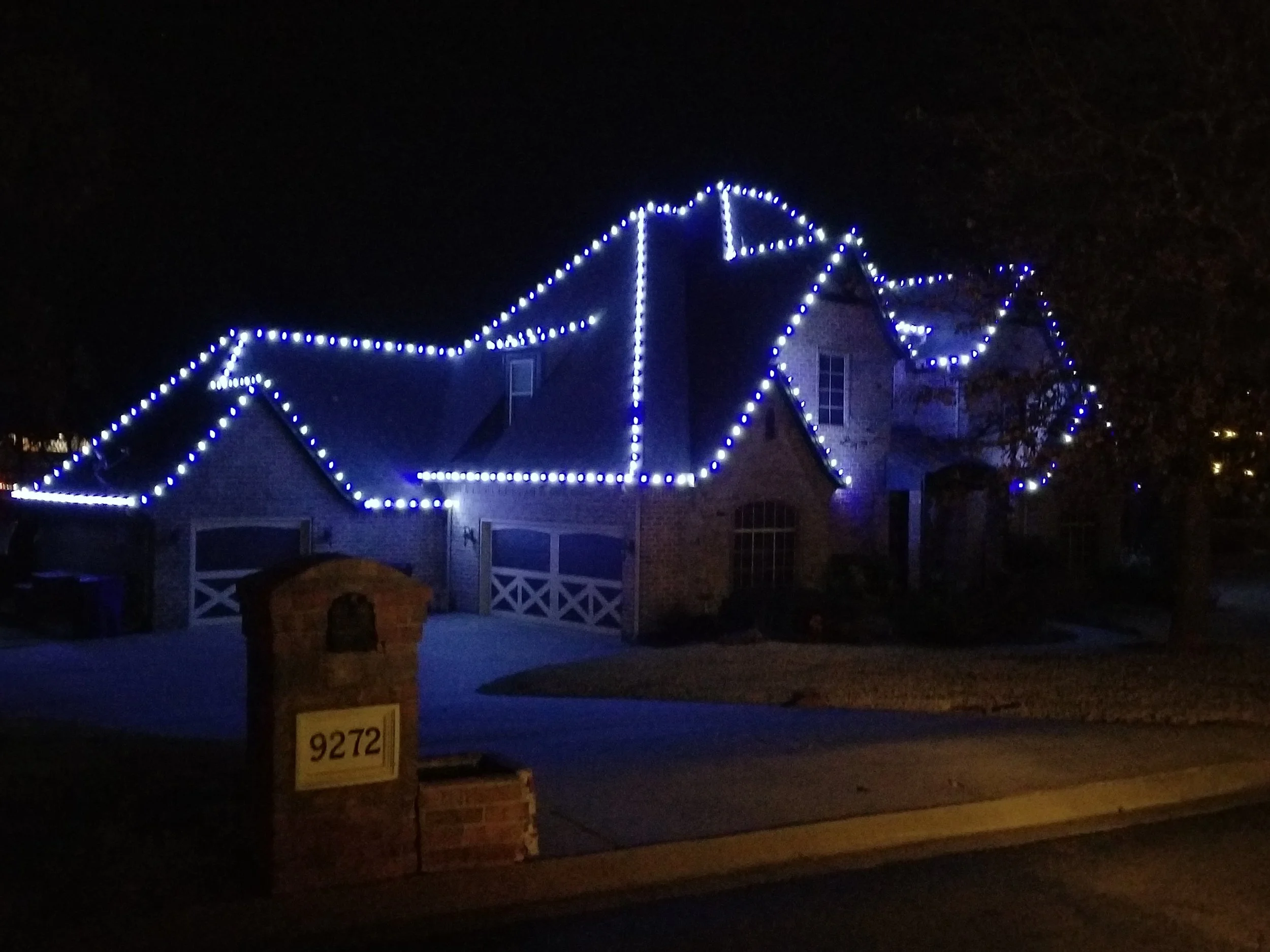 A house decorated with blue and white Christmas lights outlining the roof and windows at night.