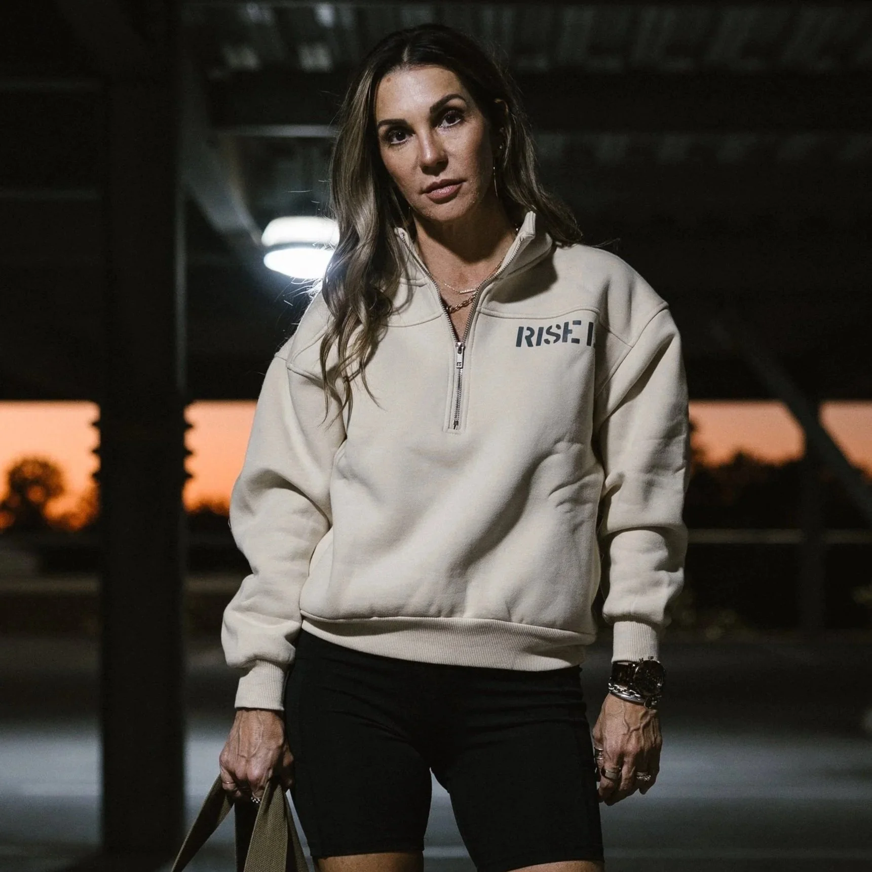 A woman standing outdoors near a parking lot during sunset, wearing a cream-colored sweatshirt with a zipper and the word 'RISK' printed on it, black shorts, and a watch, holding a tote bag.