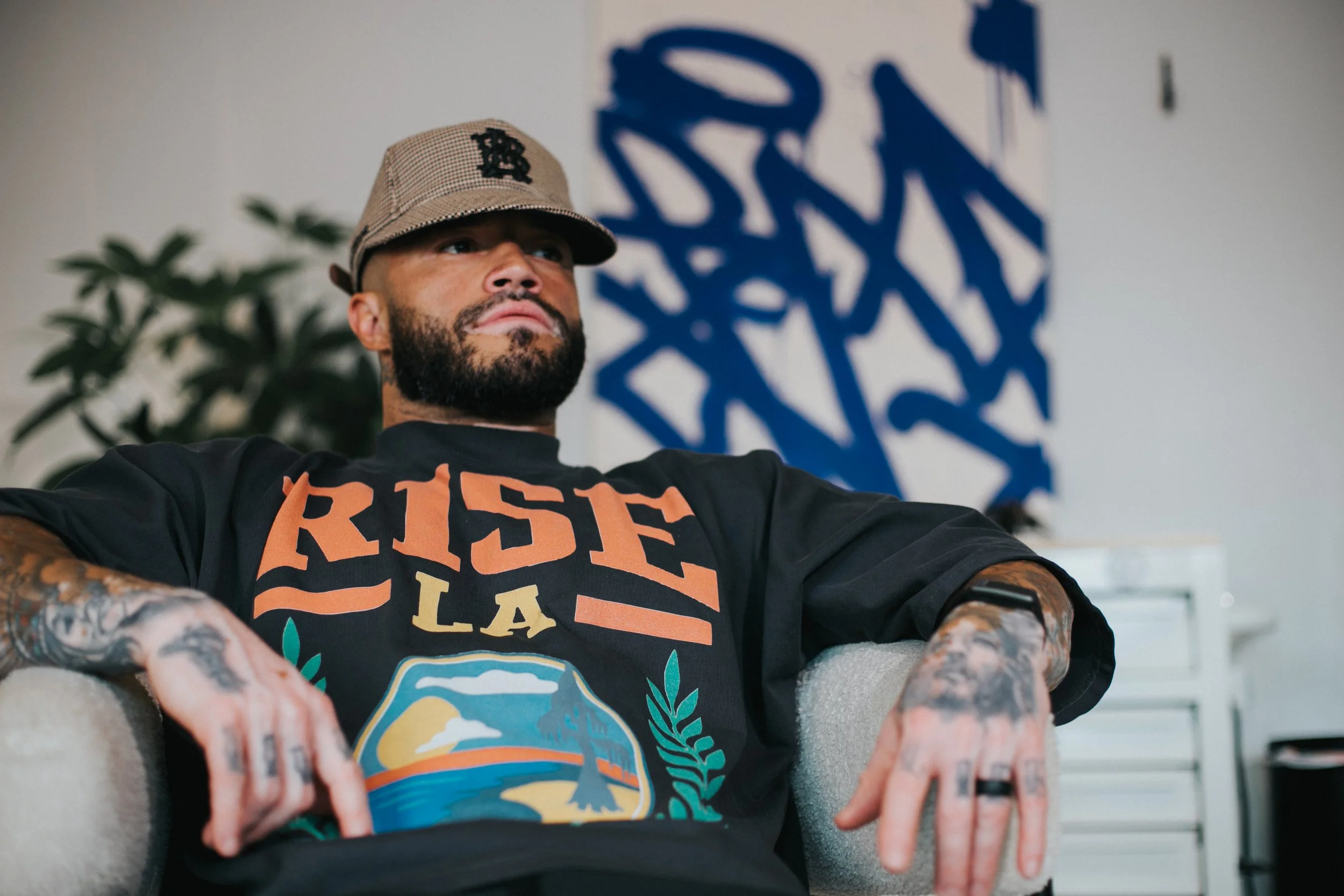 A bearded man with tattoos wearing a cap and a black T-shirt with the words "RISE LA" and a mountain design, sitting on a beige chair in a room with an abstract blue artwork on the wall behind him.