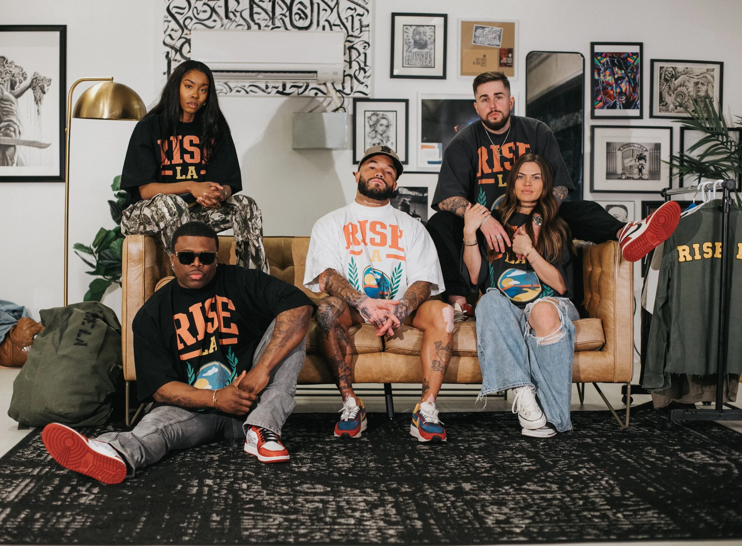 A group of five diverse young adults sitting and standing on a tan sofa in an art-filled room, wearing matching 'Rise LA' t-shirts and casual streetwear.