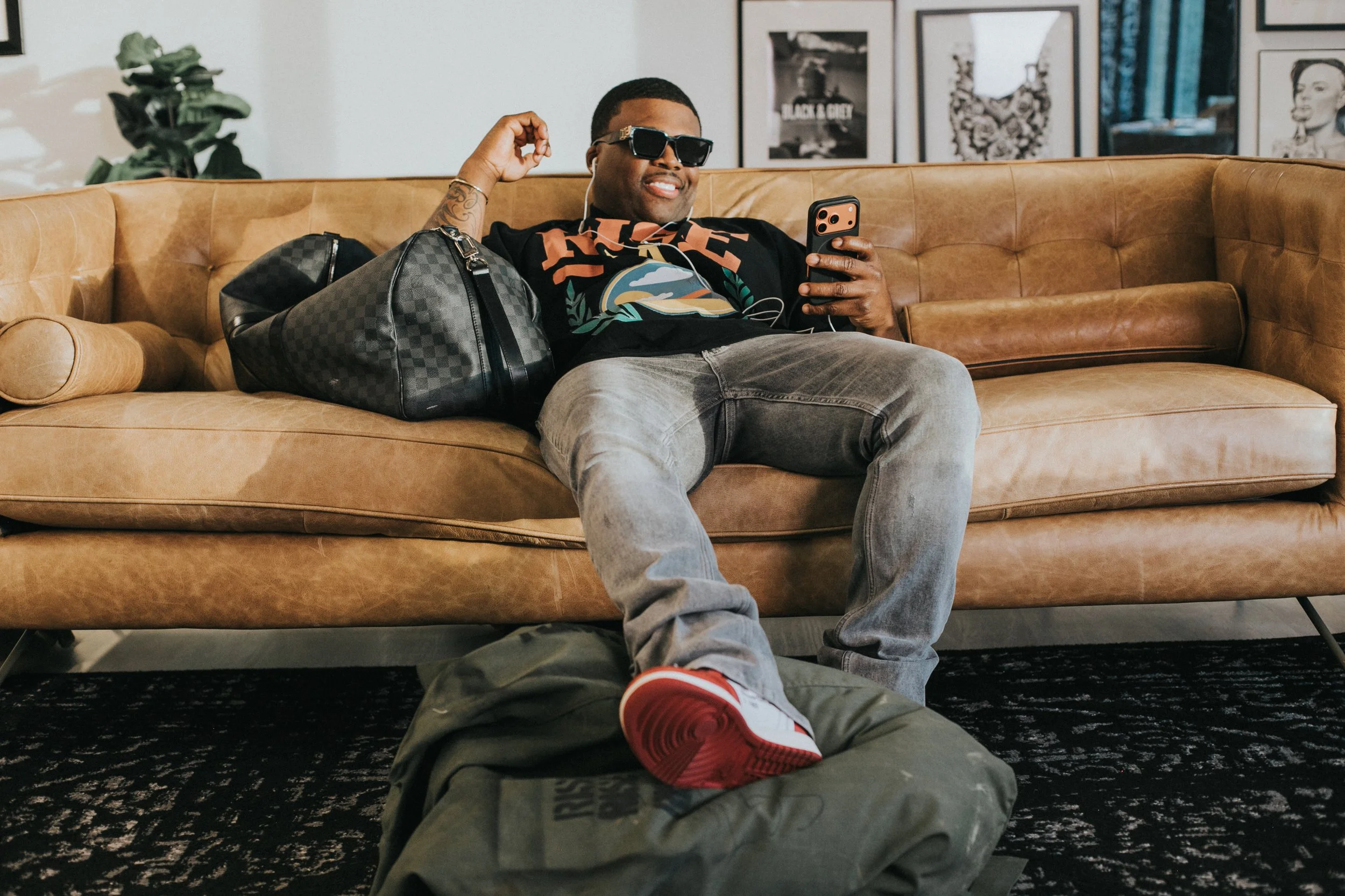 A man relaxing on a leather couch inside a modern room, wearing sunglasses, a graphic t-shirt, jeans, and red and white sneakers, holding a smartphone, with a black checkered bag beside him and art prints hanging on the wall behind.