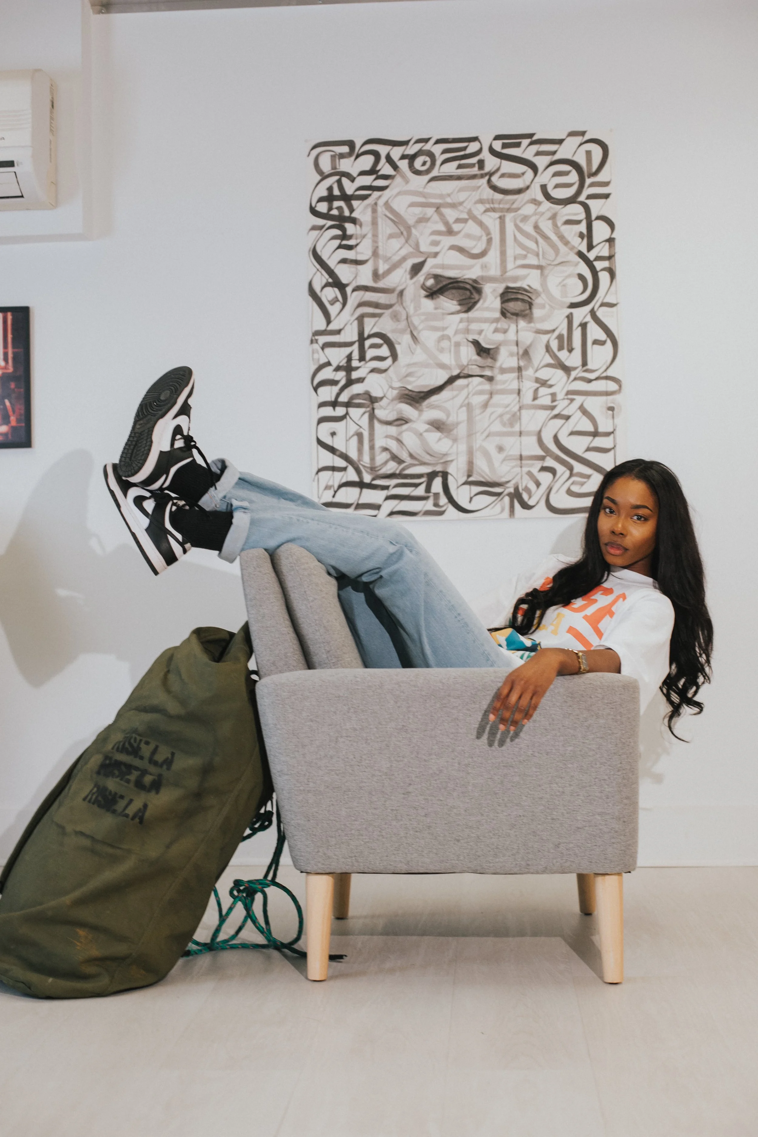 A young woman sitting in a gray armchair with her legs resting on the armrest, wearing sneakers and casual clothing, with a large green duffel bag on the floor beside her. There is a modern art piece on the white wall behind her and an air conditioni