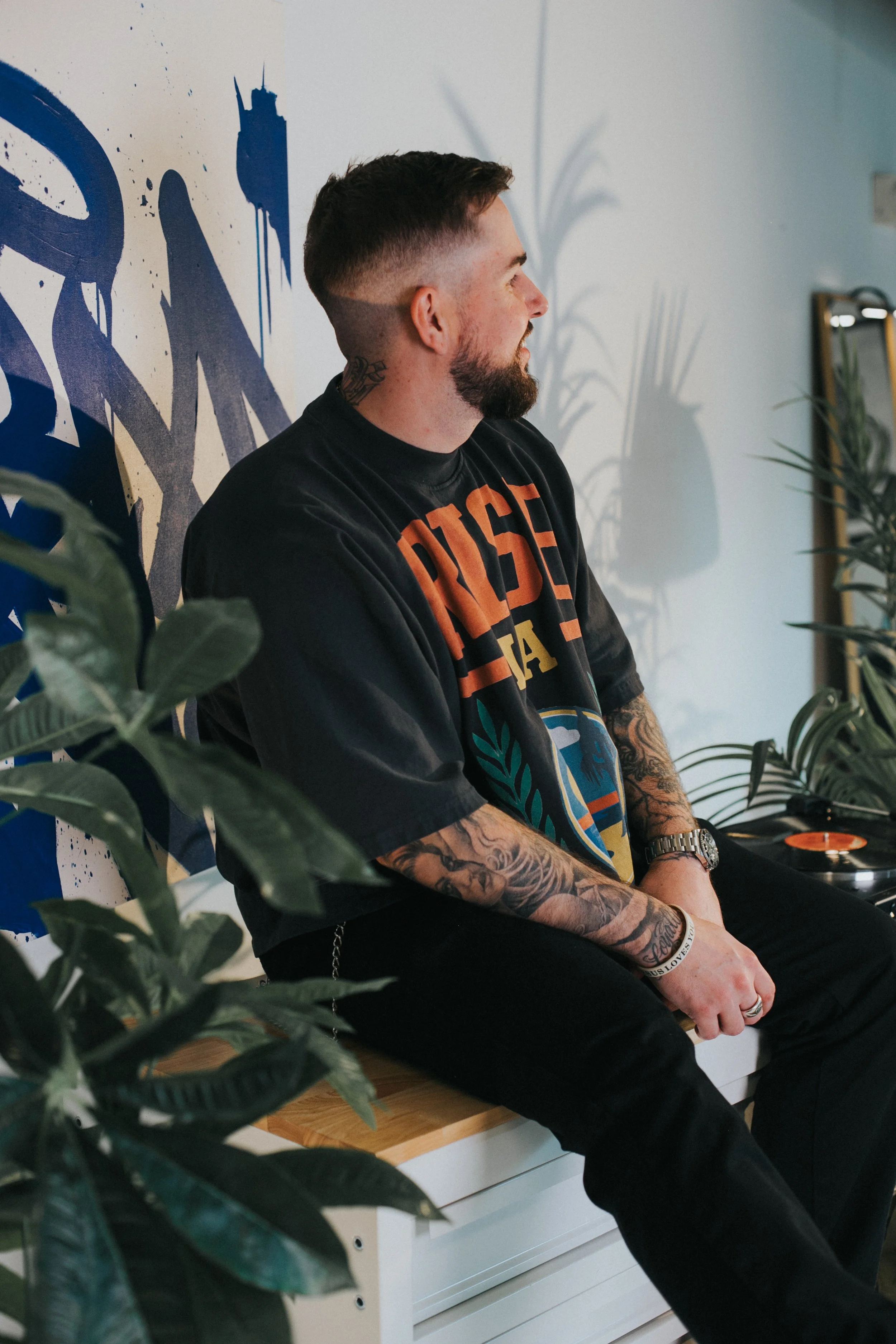 A man with tattoos on his arms, beard, and short hair, sitting on a wooden bench with a black T-shirt that has colorful text and graphic design, surrounded by indoor plants, looking to the right.