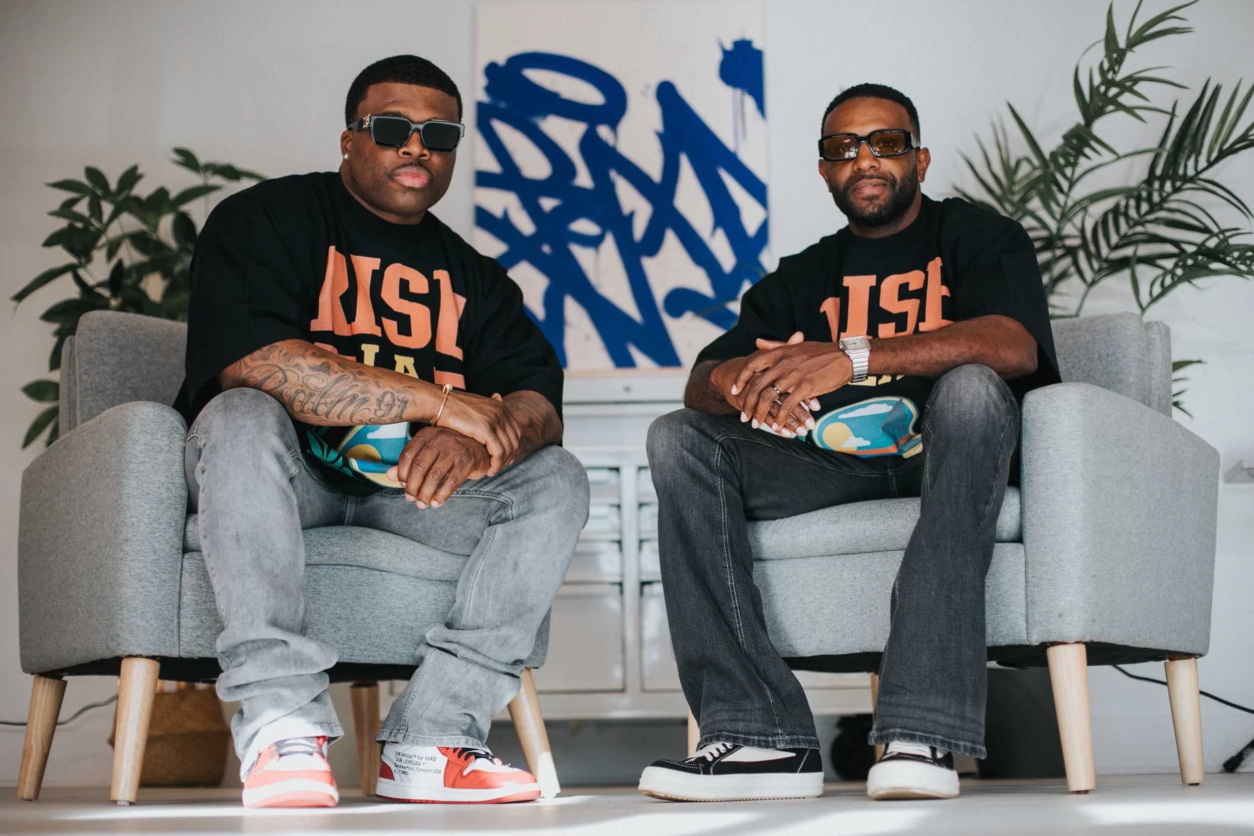 Two men sitting on gray armchairs indoors, wearing black T-shirts with orange lettering, sunglasses, and sneakers. There are green plants and an abstract art piece with blue brushstrokes in the background.