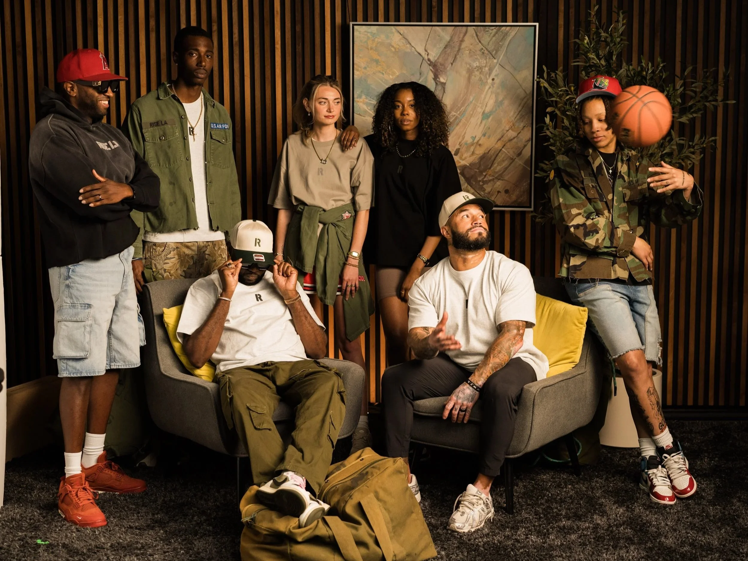 Group of eight young adults, some sitting, some standing, in a modern room with wooden wall paneling and a painting, with one person spinning a basketball on their finger.
