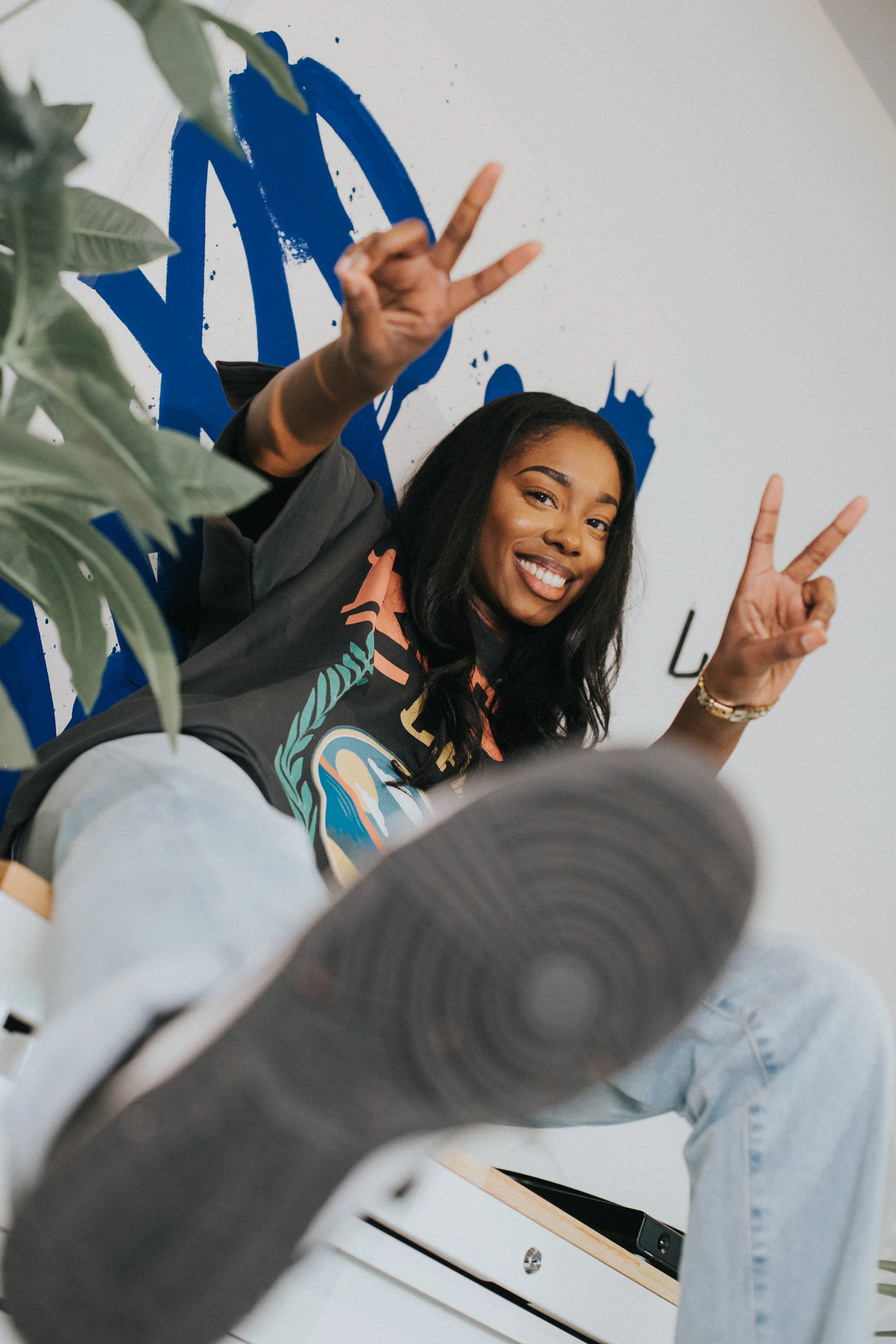 Smiling young woman sitting on a white bench, making peace signs with both hands, with a blue graphic background and some greenery in the foreground.