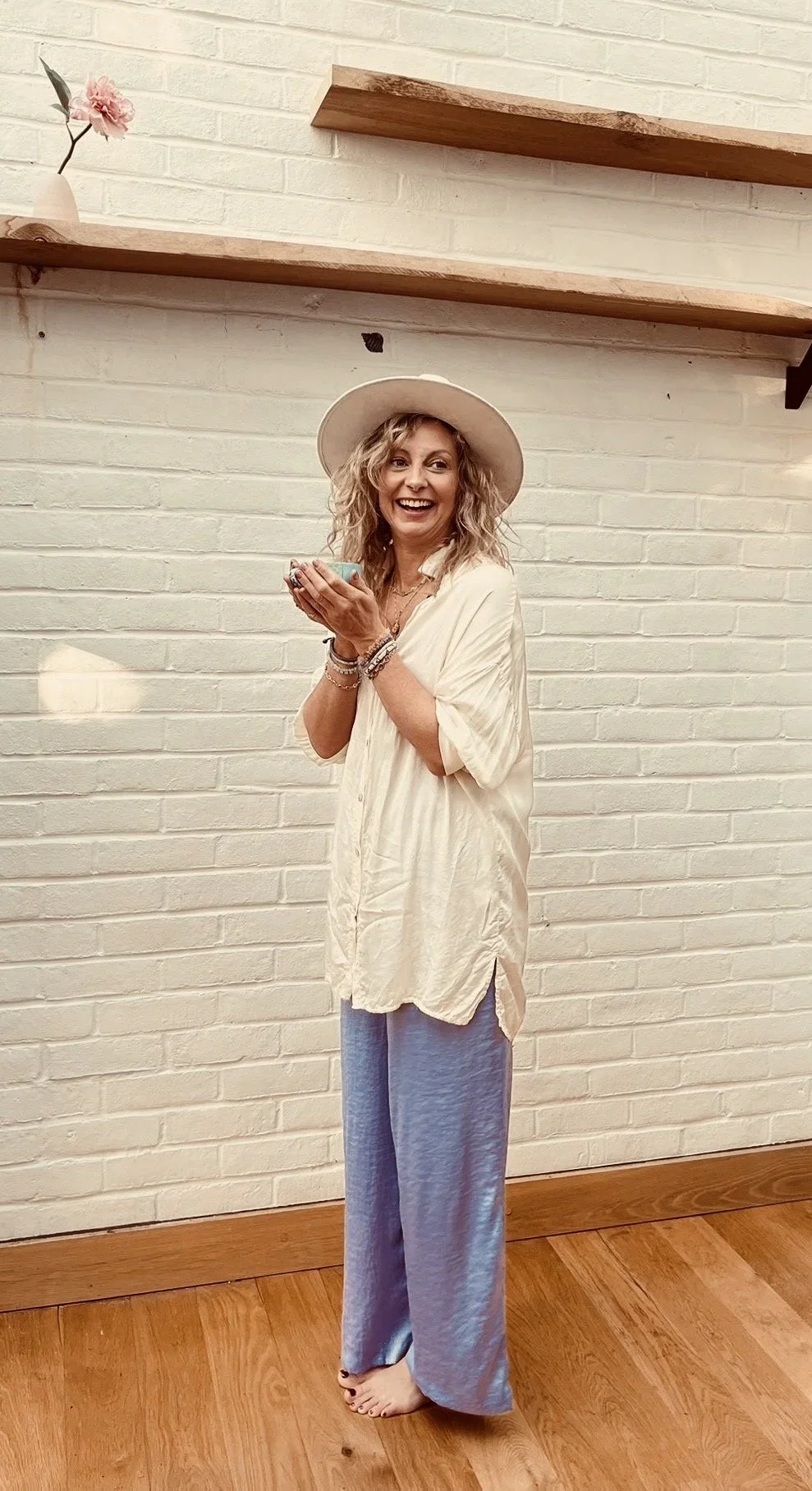 A woman with curly blonde hair, pink tassel earrings, and a blue animal print top smiling inside a cafe with large windows and hanging white lights.