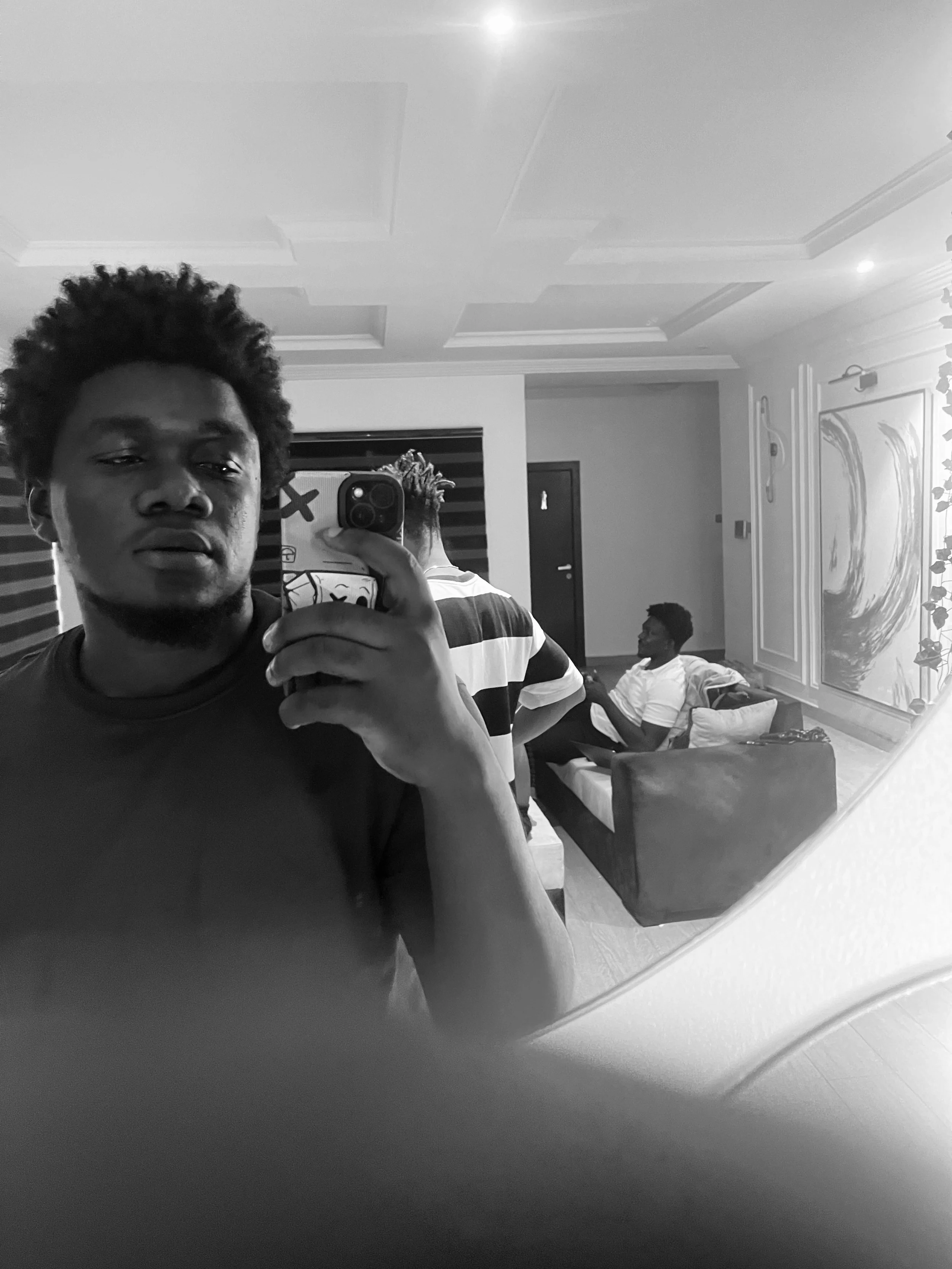 Black and white photo of a man taking a mirror selfie with a smartphone in a room. Another person is sitting on a couch in the background.
