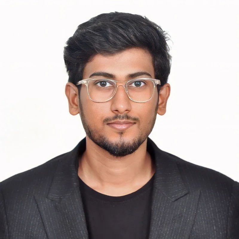 A man with glasses wearing a black shirt and blazer against a white background.