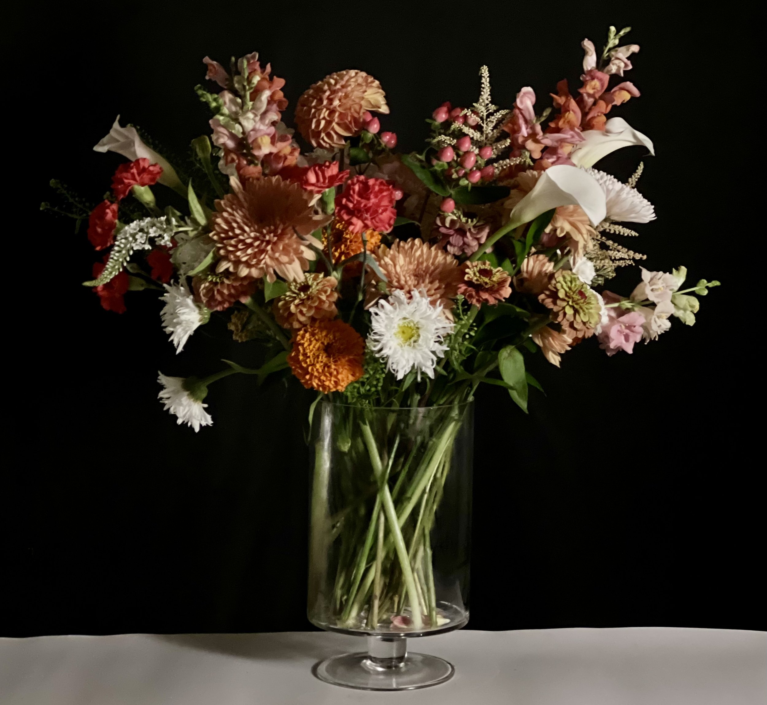 An autumn mix of blooms in a vase showing blush, reds & oranges with accents of white dahlia and calla lilies