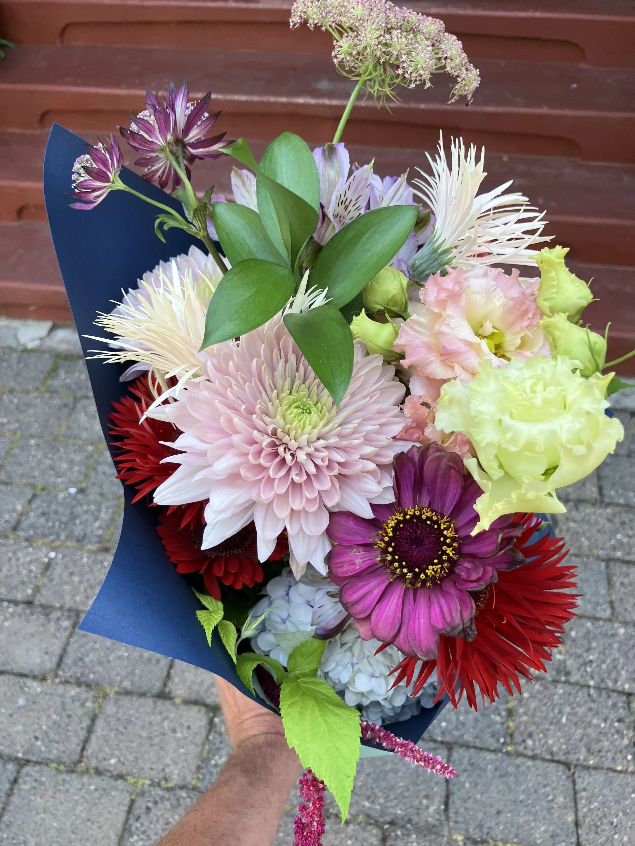 A handheld bouquet shown against a brick wall, colored with pinks, reds and yellow blooms