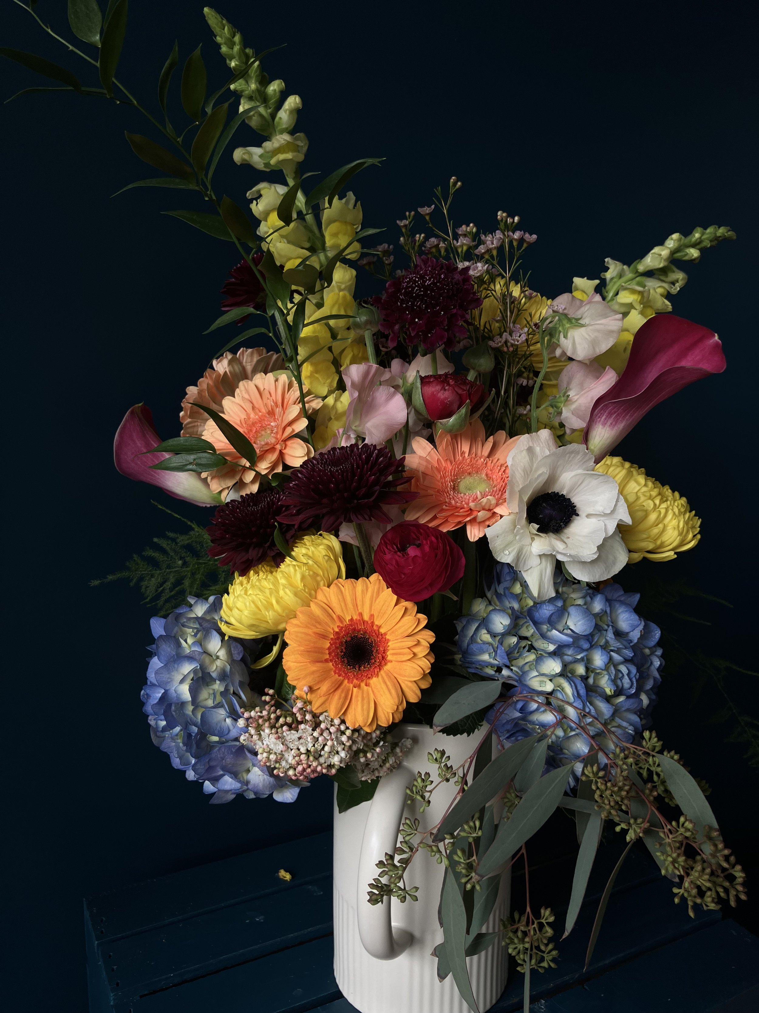 Blue hydrangeas surrounded by a colorful combination of anemone, gerbera daisy & chrysanthemum blooms poised in a container