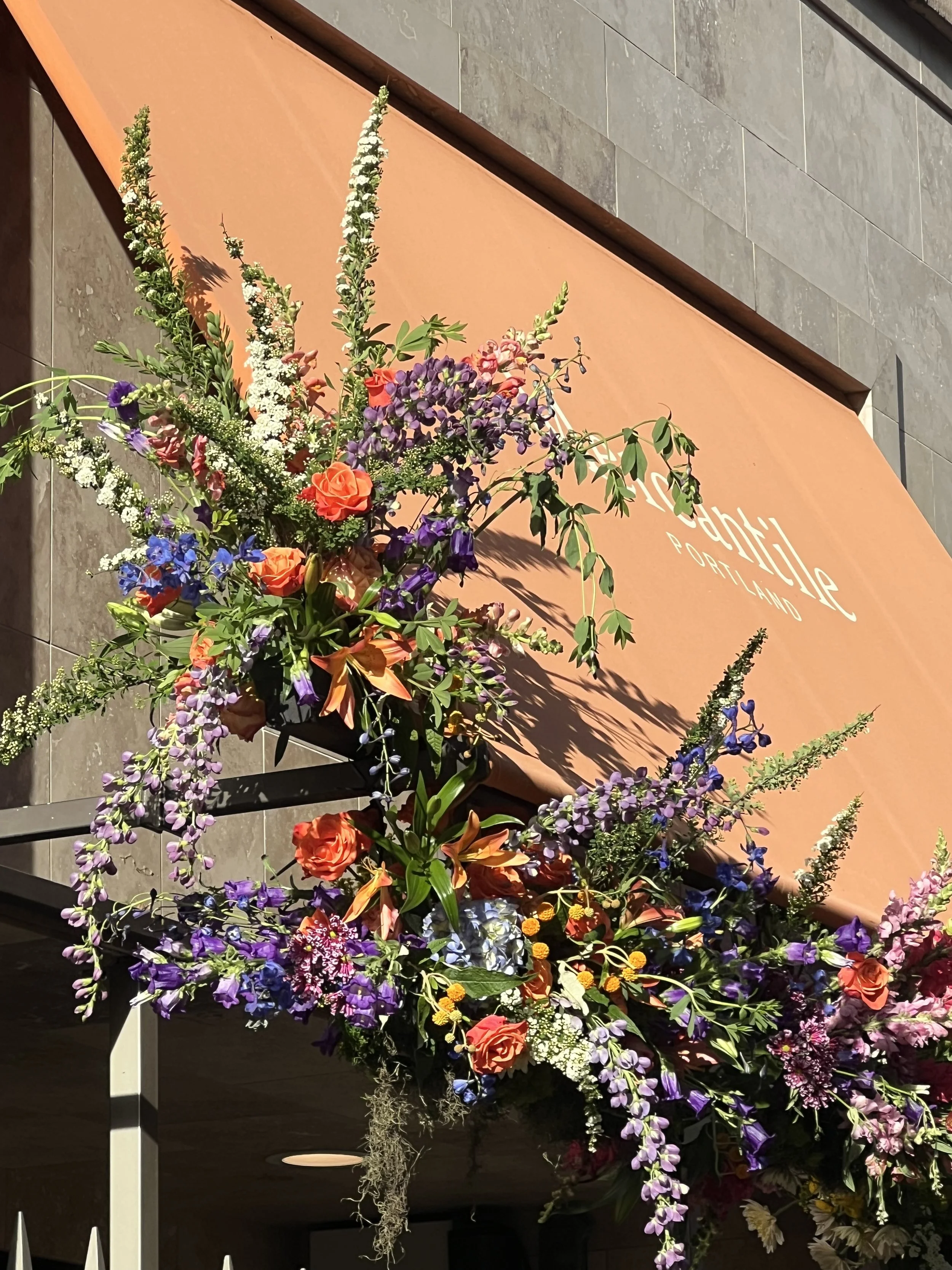A vibrant hanging floral installation with roses, lilies, delphinium, spirea and indigo, designed for a downtown Portland storefront