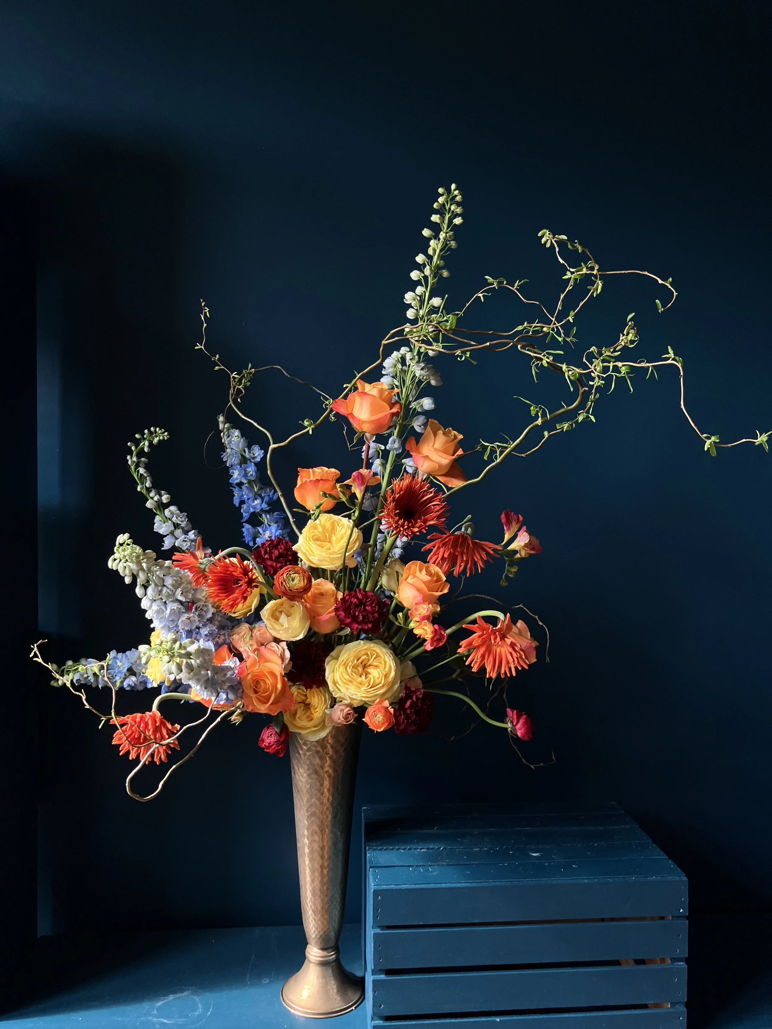 Tall brass vase of red roses, ranunculus, and blue delphinium in an asymmetrical luxury floral arrangement with rich texture and movement, made as event flowers by our Portland florist