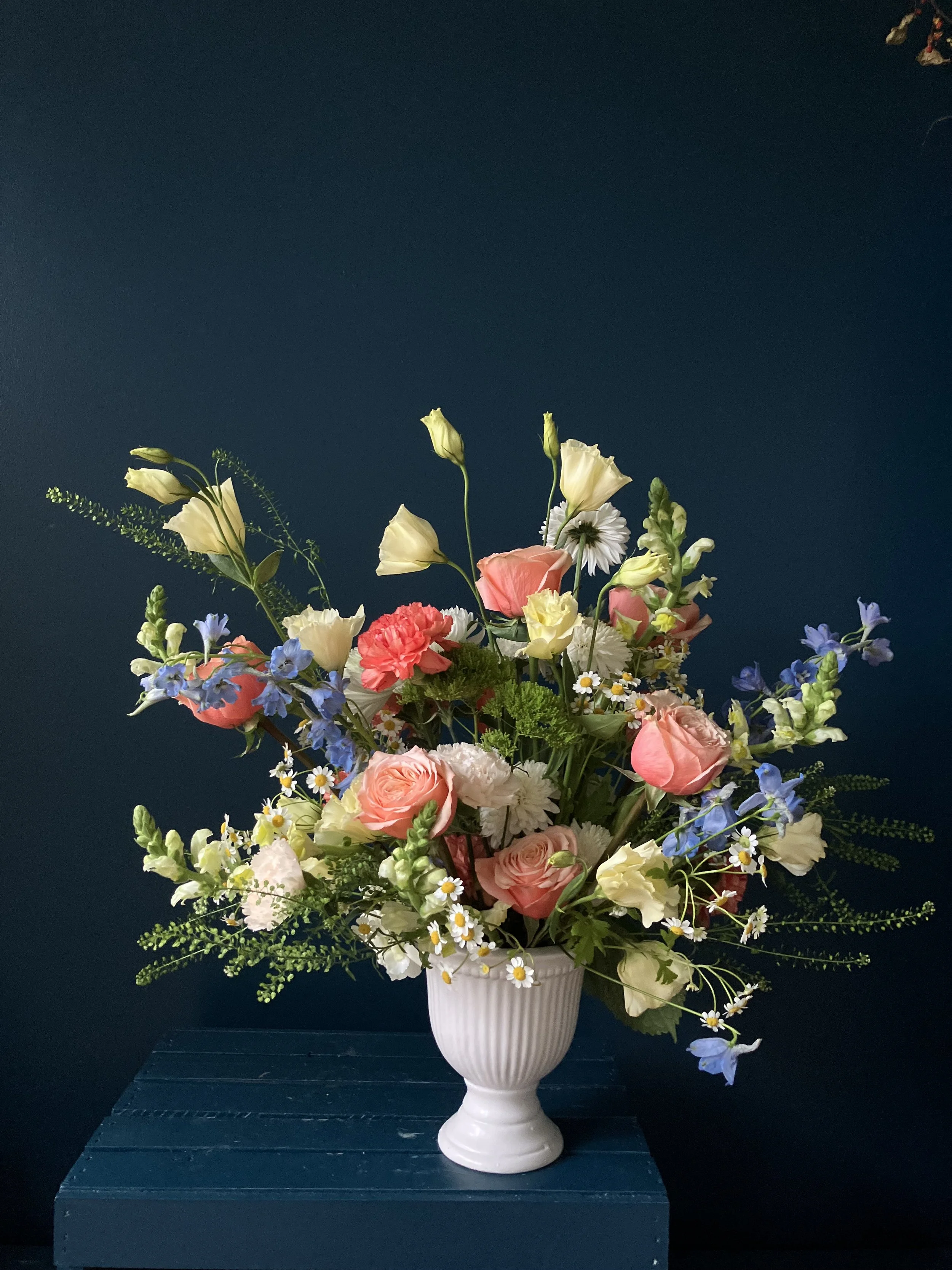 A sunnier version of our sympathy arrangement in white vase, featuring added yellow lisianthus and blue delphinium