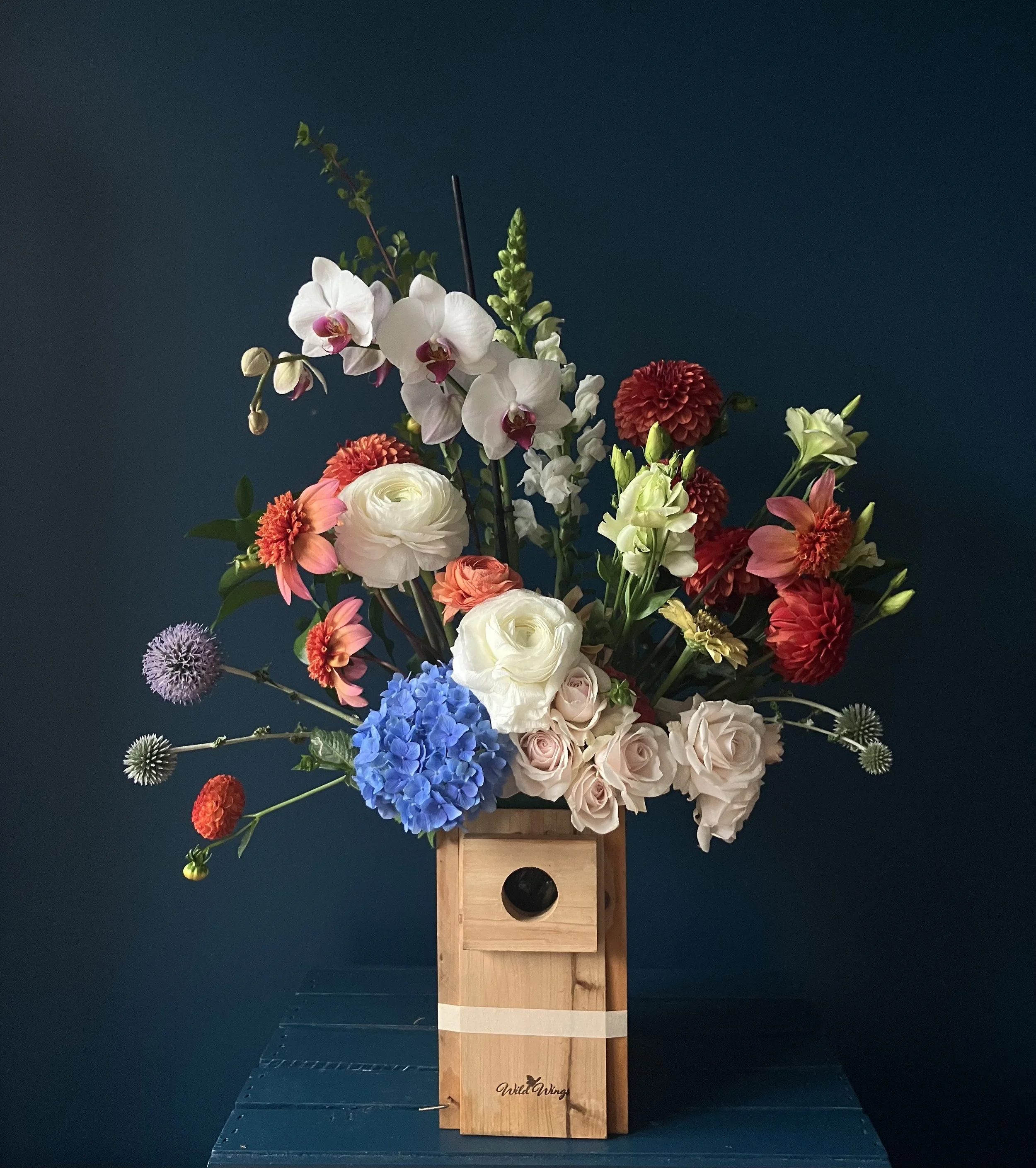Seasonal mixed version of this Portland florist arrangement in bird box with ranunculus, dahlia, rose and a striking orchid