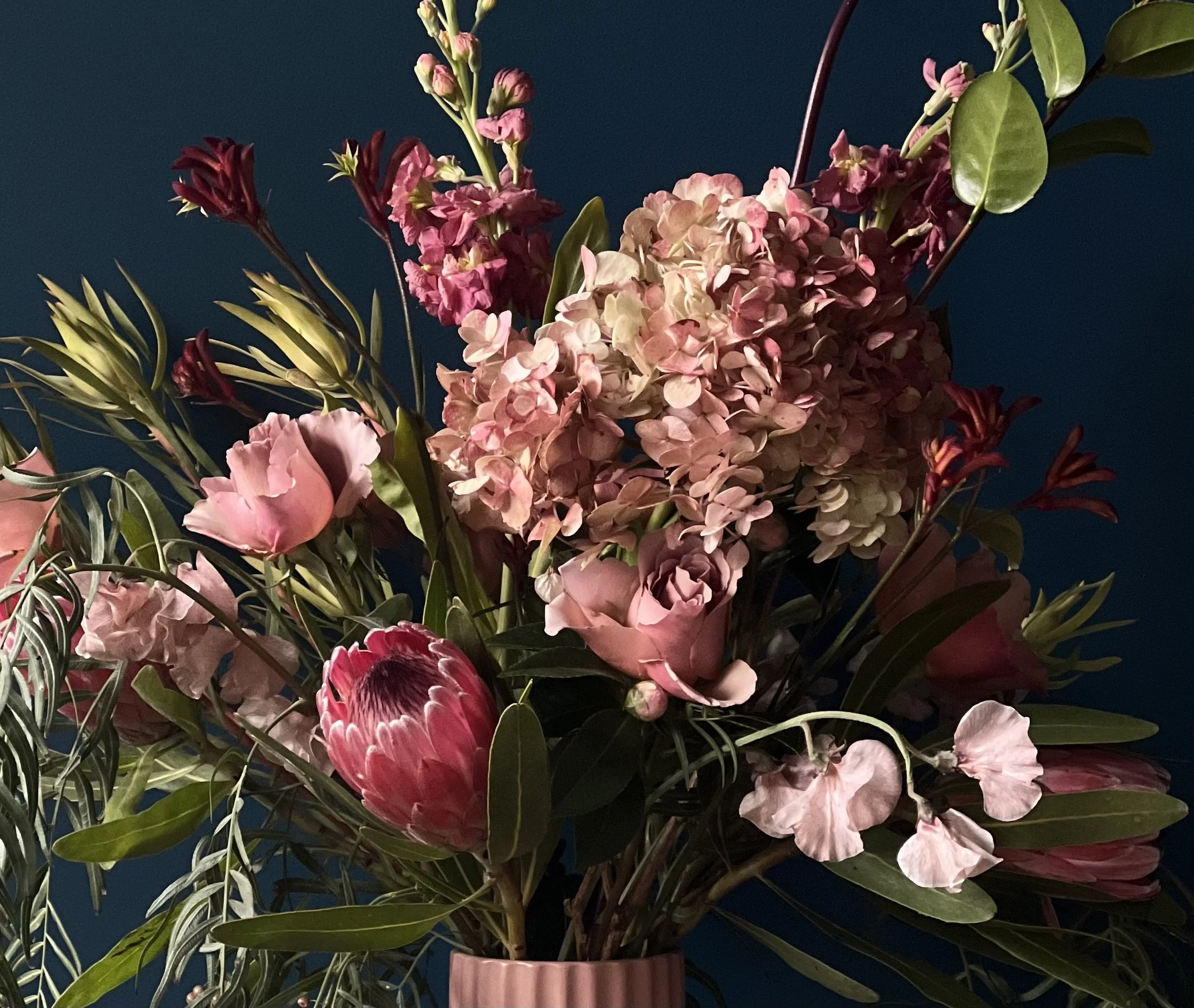 Close up view of blush-toned roses. soft pea blooms, and ruddy hydrangea in an arrangement with tropical protea and foliage