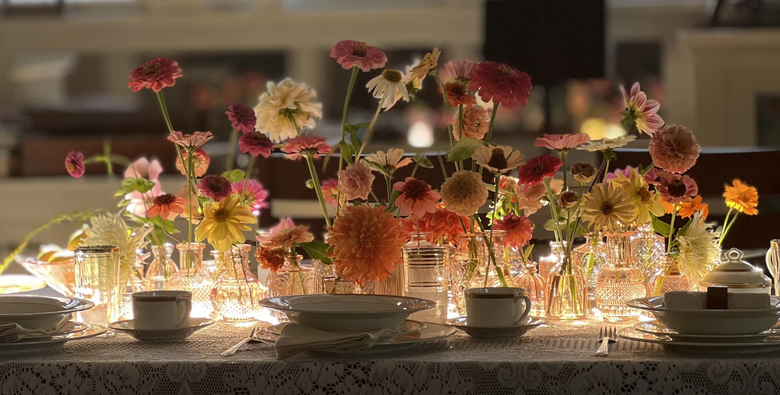A candlelit wedding table with dinnerware and dahlia and zinnia blooms in bud vases