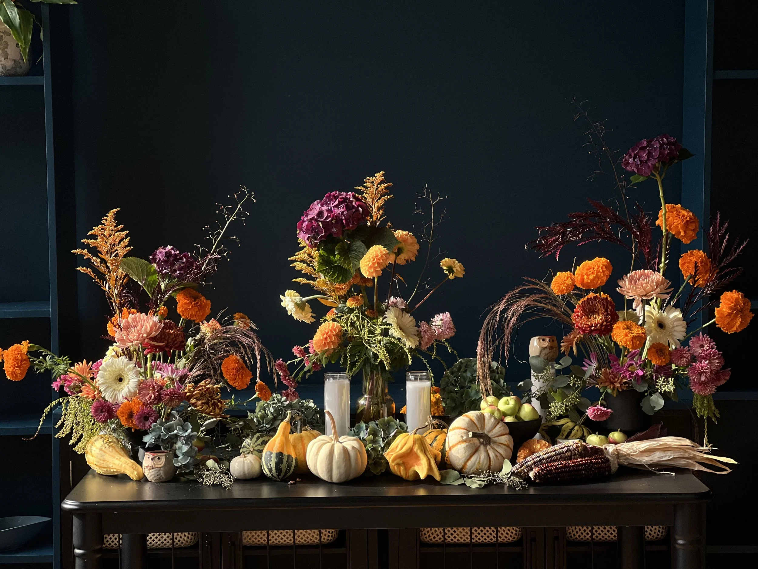 Autumnal floral arrangement with pumpkins, gourds, and candles on a dark table against a dark wall.