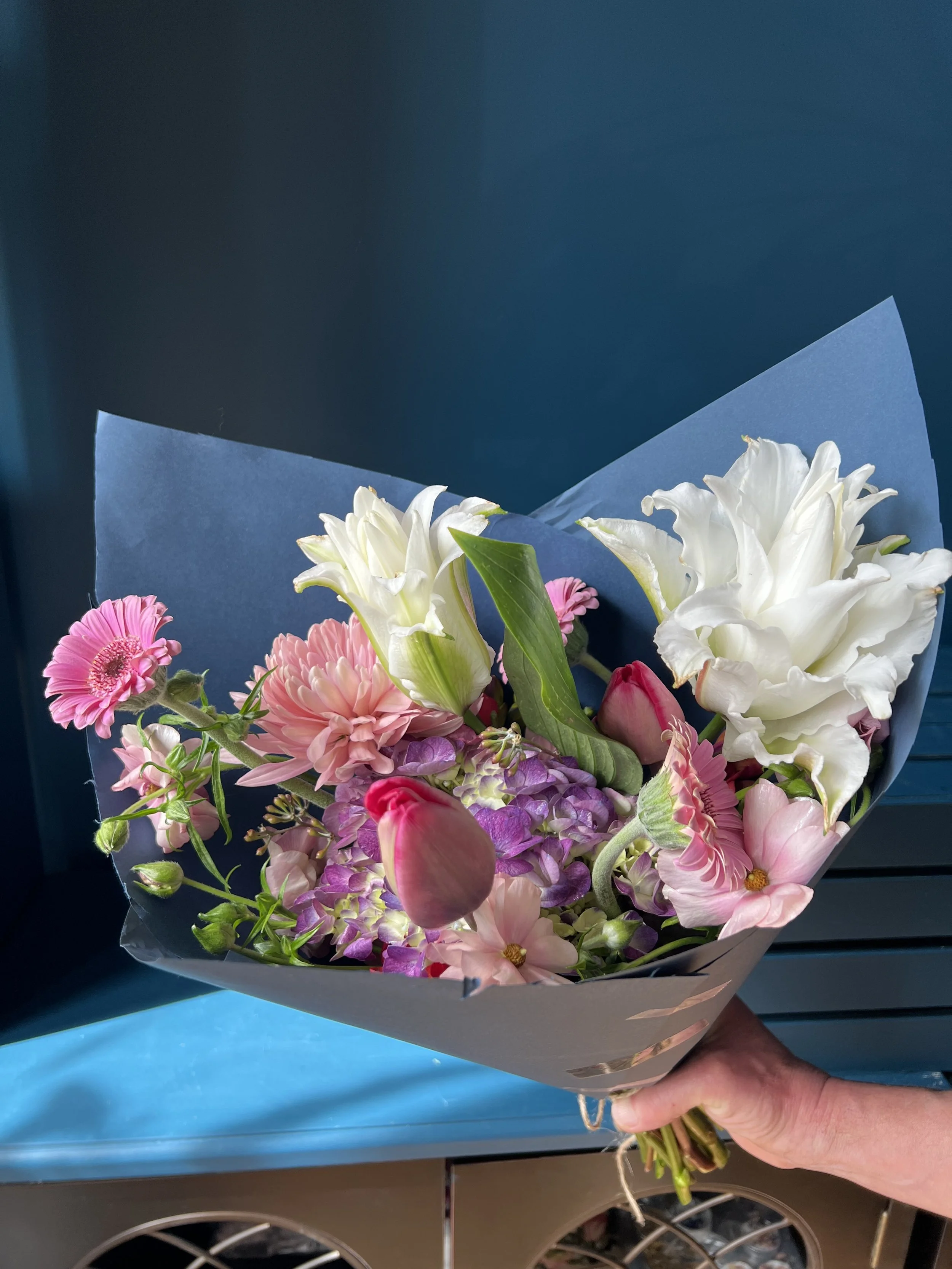A spring seasonal bouquet with white lilies, callas, blush mum, and unopened red tulips, handheld against blue backdrop