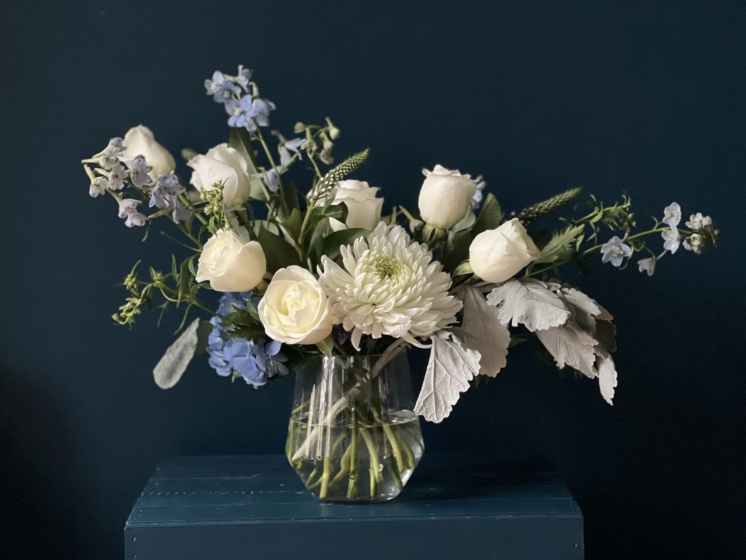 Lush winter floral arrangement in Portland Oregon featuring white roses, mums, blue delphinium and hydrangea in a clear vase against a dark background
