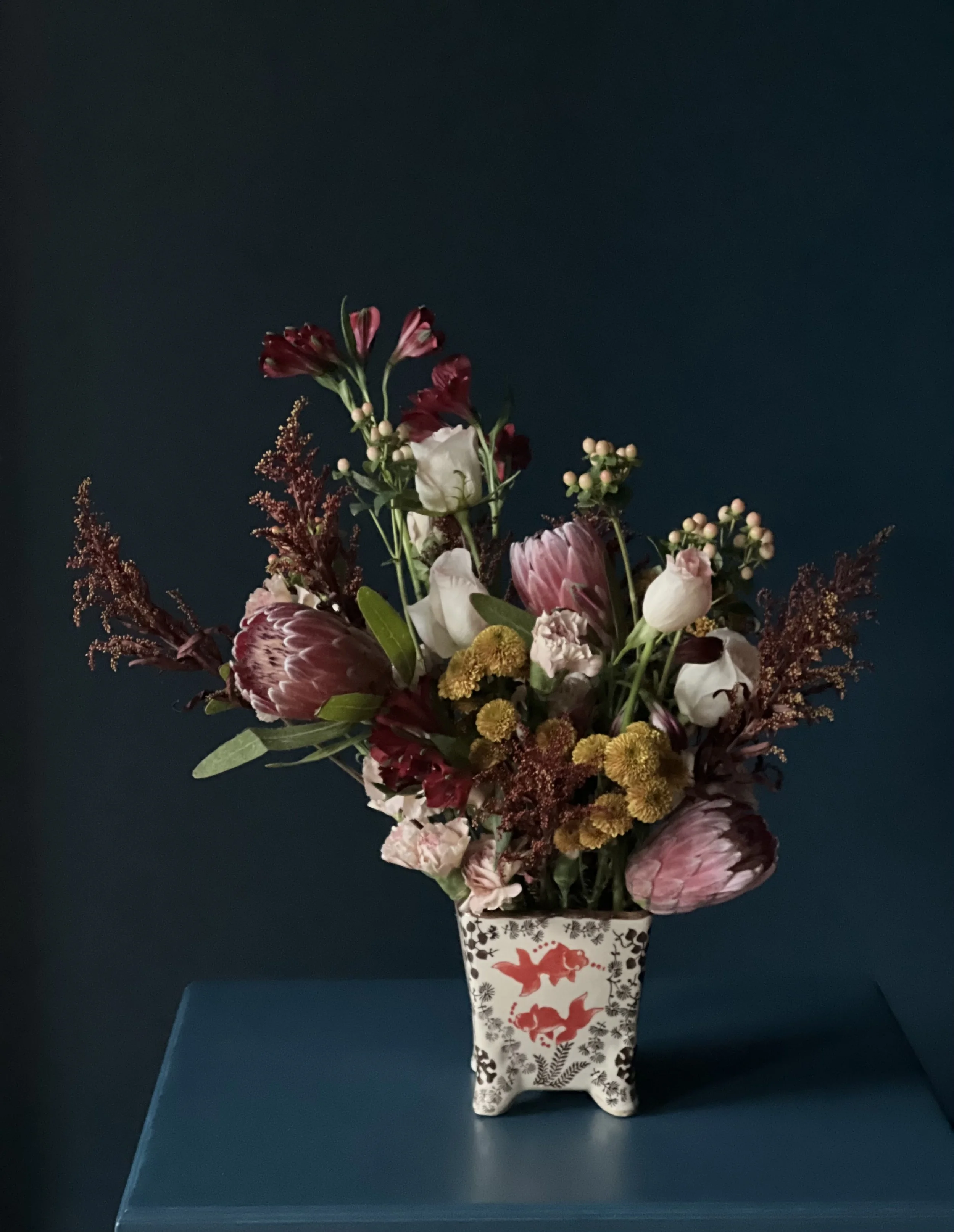 Textural semi-tropical floral arrangement with red protea, roses white, and fillers in a dynamic, garden-style design.