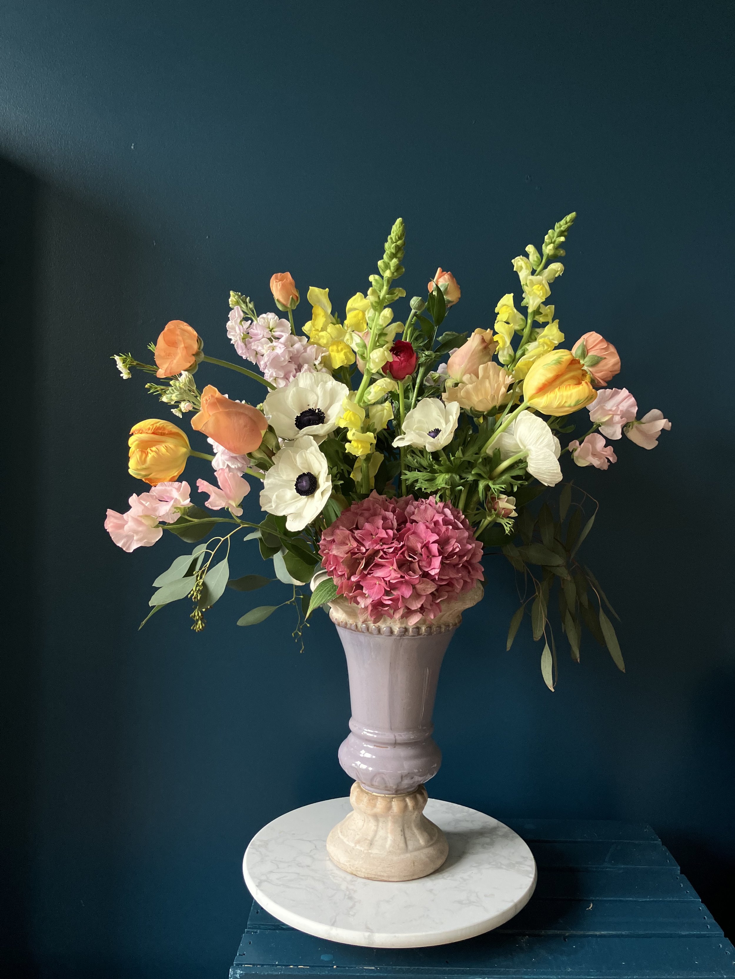 Multi-hued anemones, red ranunculus, yellow snapdragons & more flowers grace an urn in this spring bouquet gift