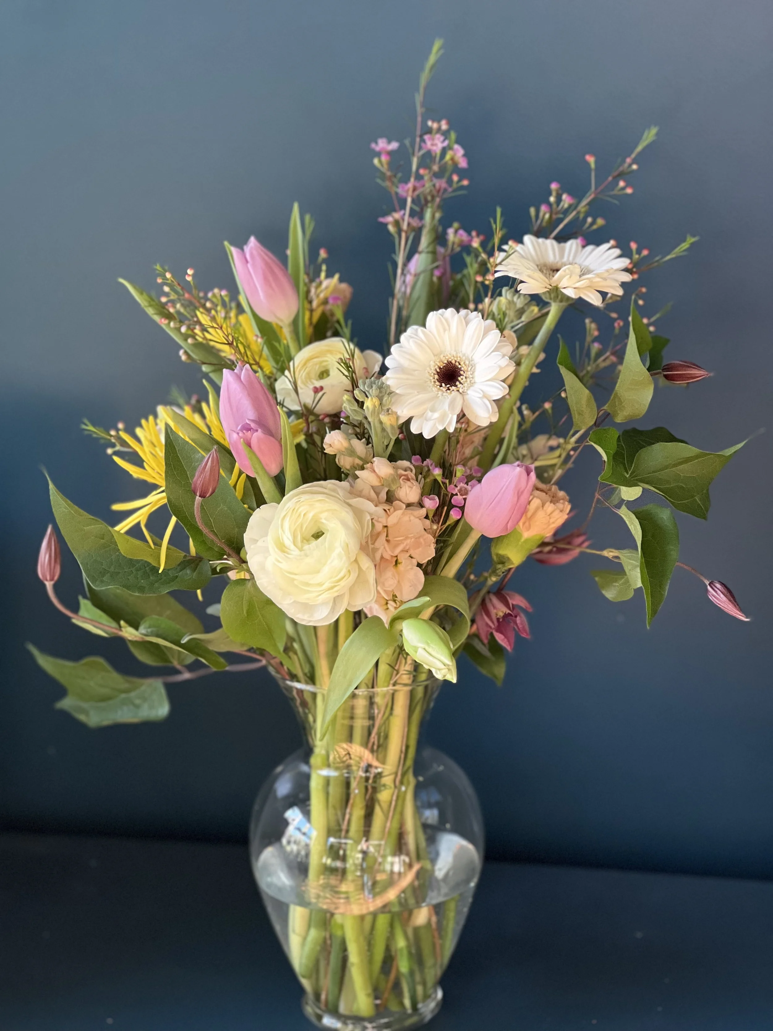 Held Close sympathy bouquet with white ranunculus, lavender tulips, yellow mums, and daisies in a glass vase, ready for Portland hospital delivery
