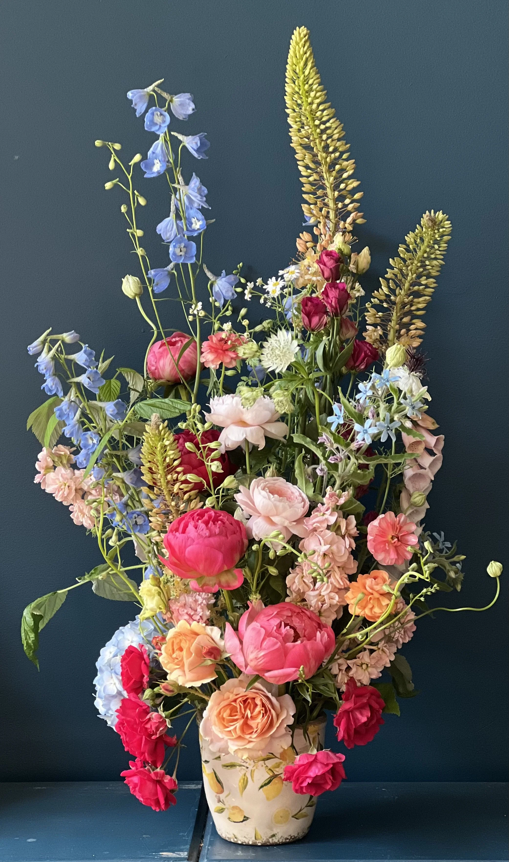 Moving floral column in a burgundy-peach color scheme in a vase, accented with delphinium and eremerus, designed as a garden party centerpiece