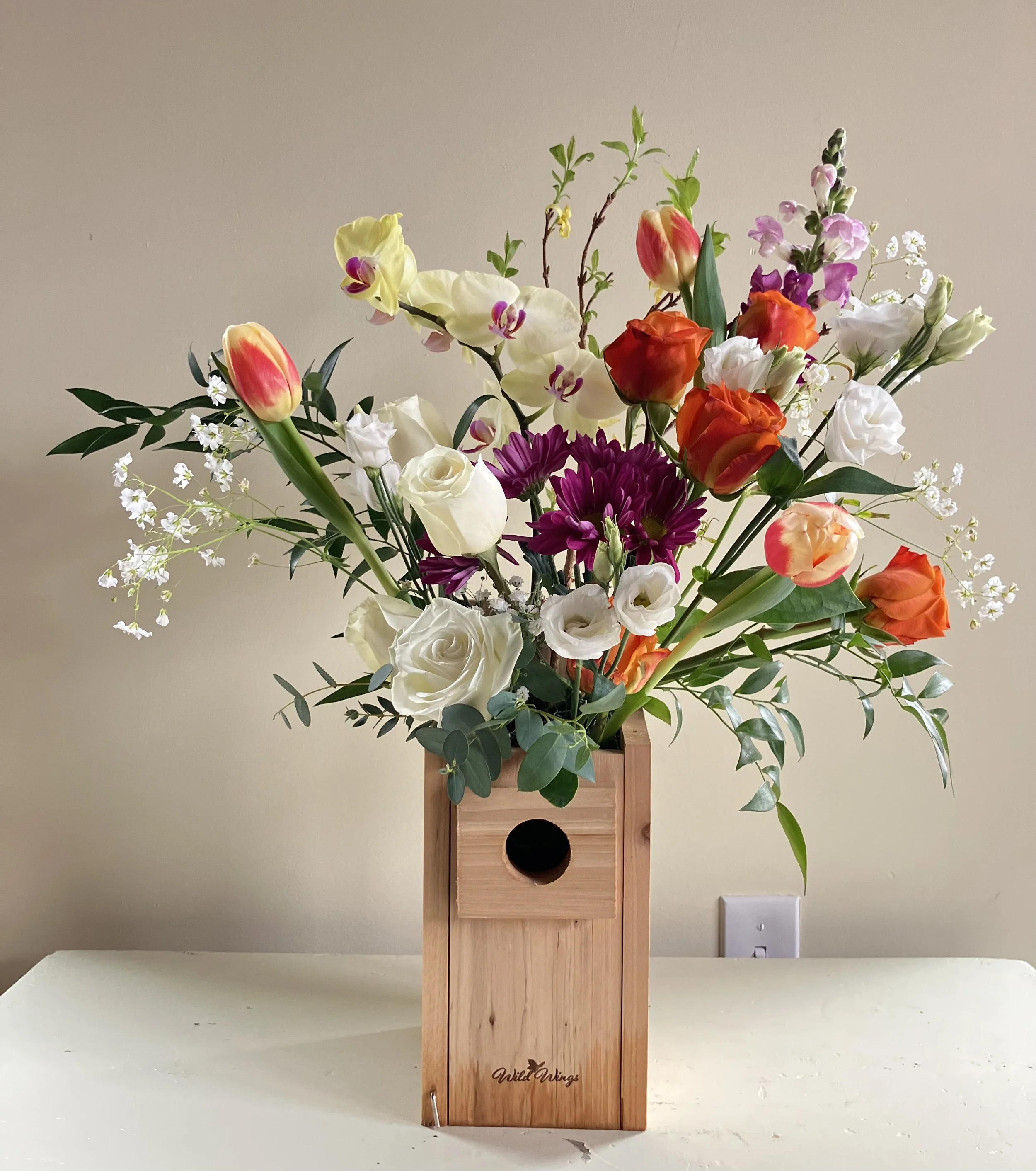 White & orange roses, tulips, purple pea and an arcing orchid perched in a bird box, designed as a Portland floral gift