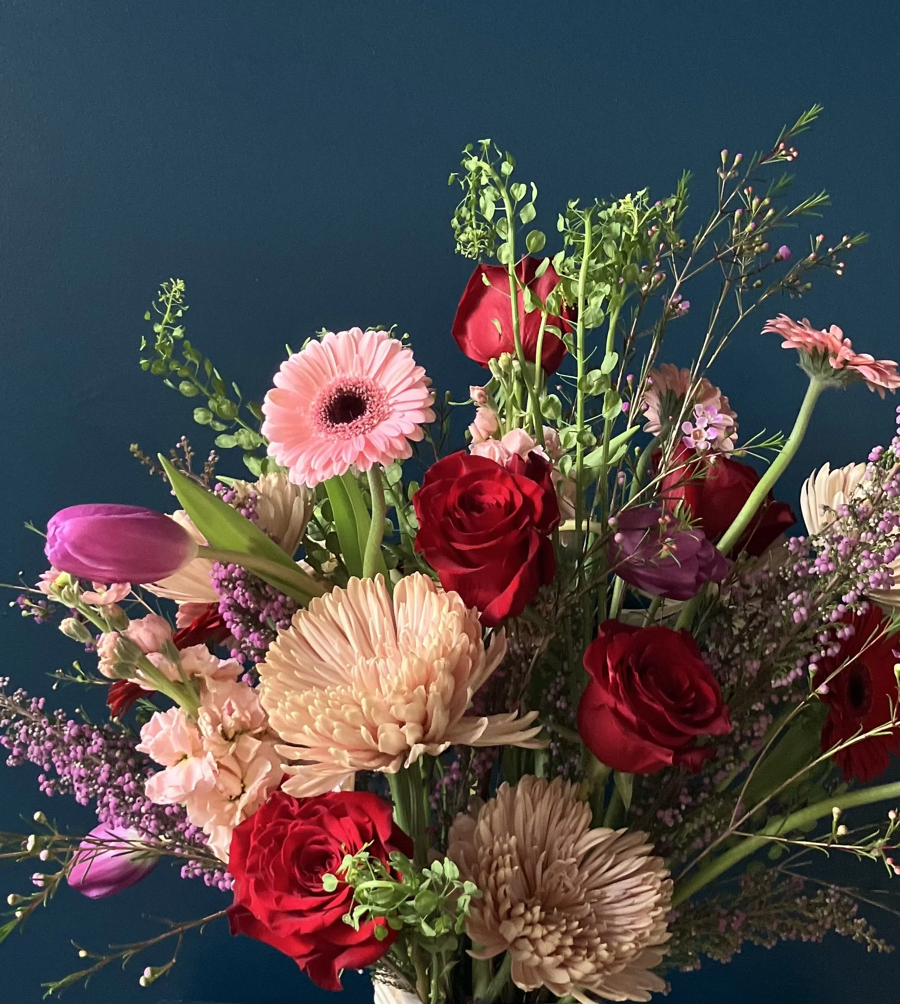 Close-up of red roses, pink stock, light purple tulips, and textured foliage in a Valentine’s arrangement, highlighting sculptural floral design and movement.