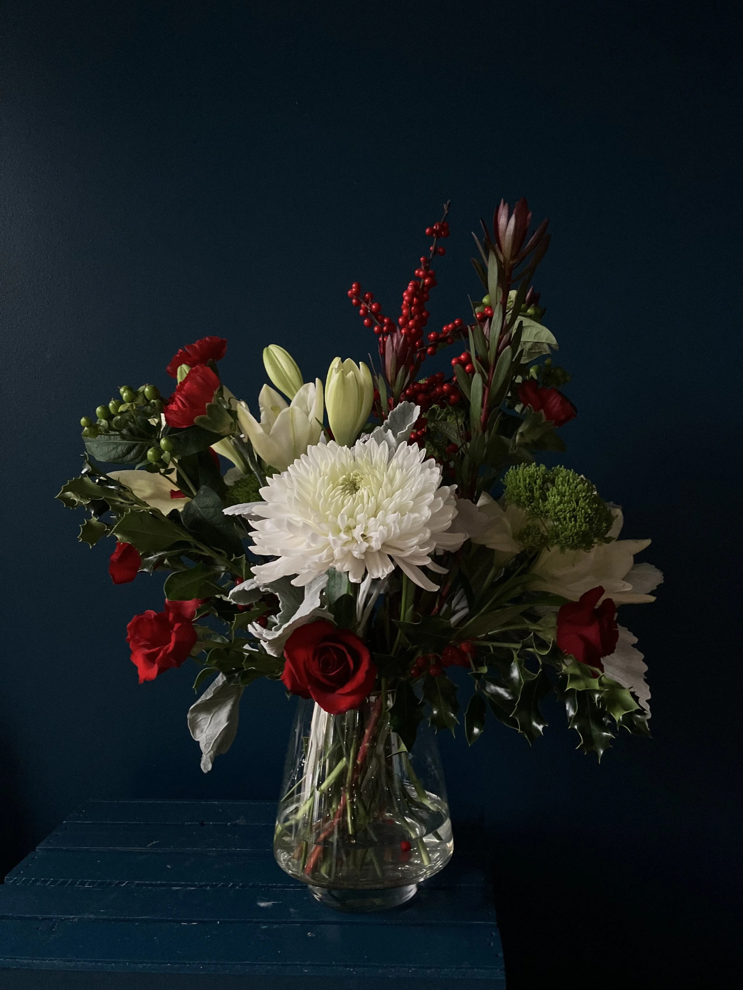 A Christmas bouquet in a clear vessel with local and non-local blooms of red roses, white lilies and carnation filled out with foliage