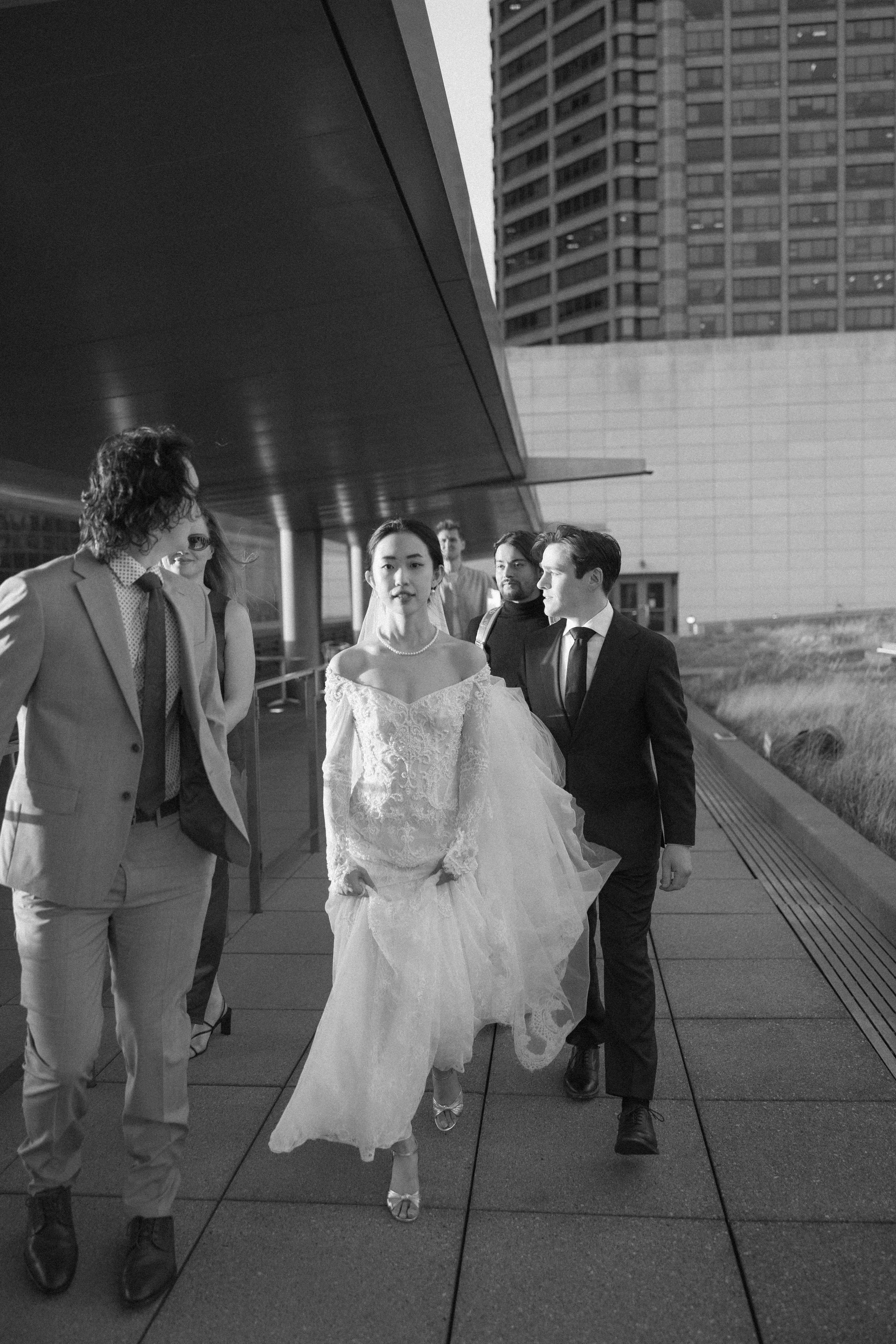 A group of people, including a woman in a wedding dress, walking together outside in an urban setting with tall buildings.