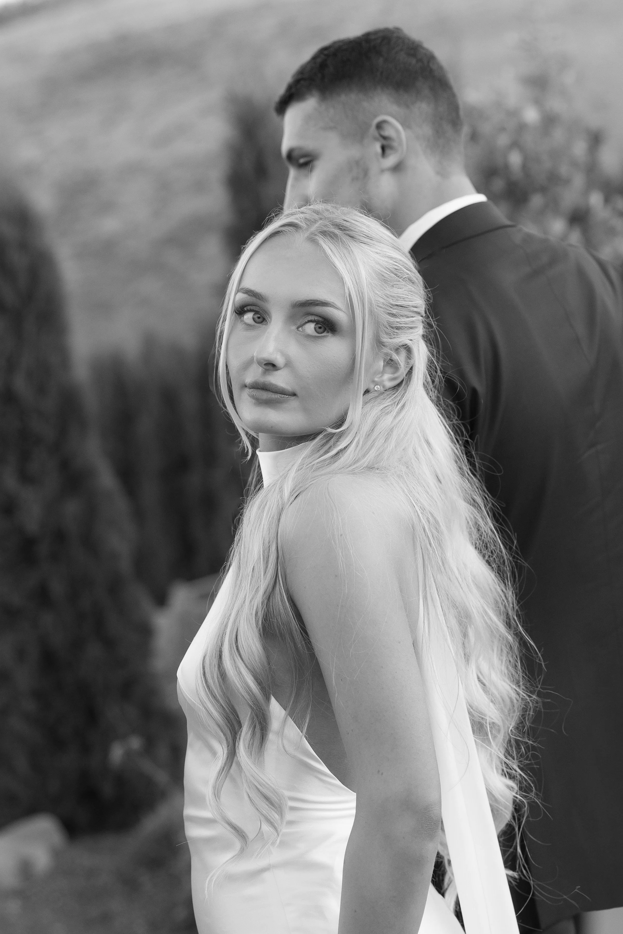 Black and white photo of a woman with long blonde hair and a man in a suit standing behind her outdoors.