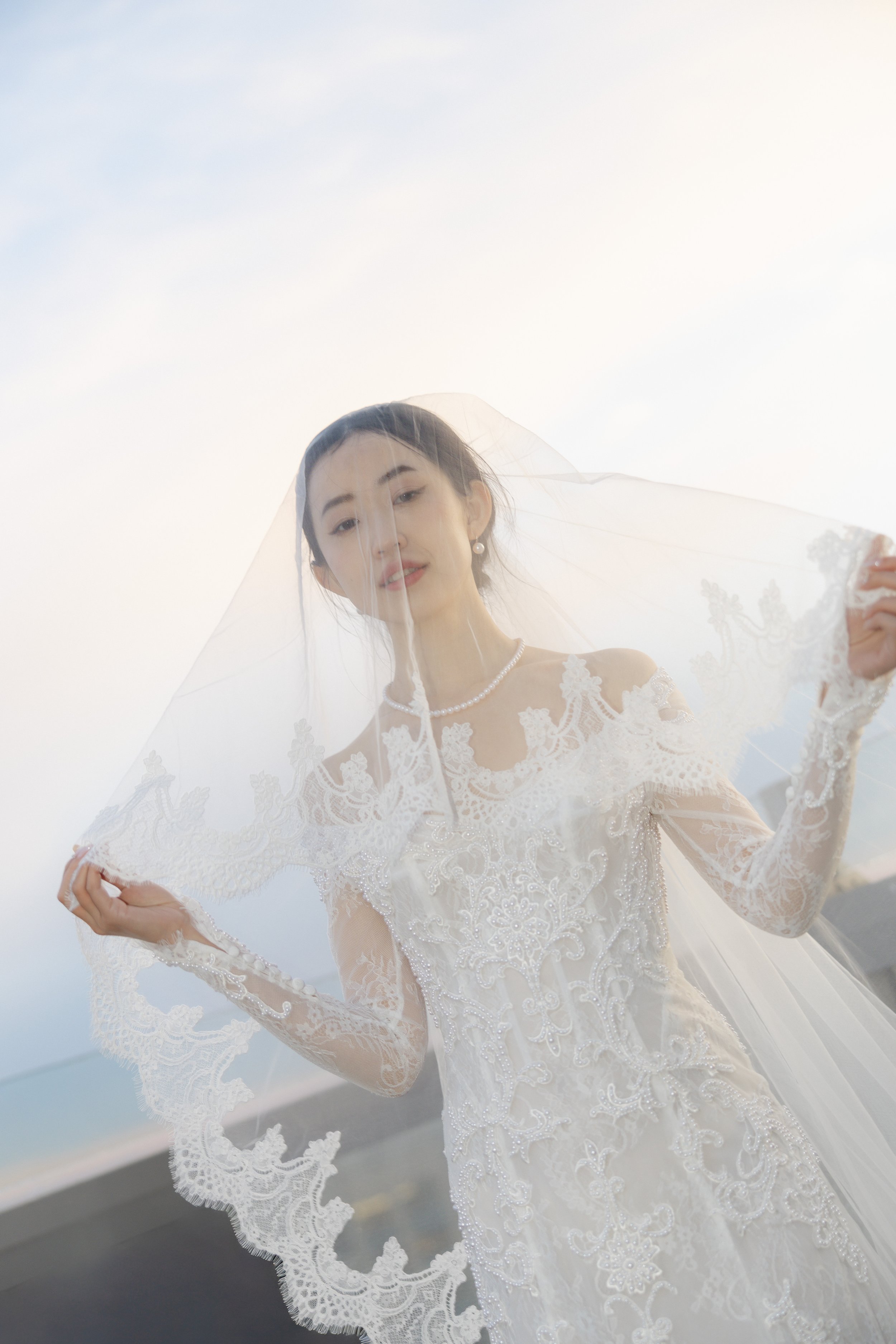 A bride in a lace wedding dress holding a veil outdoors against a bright sky.