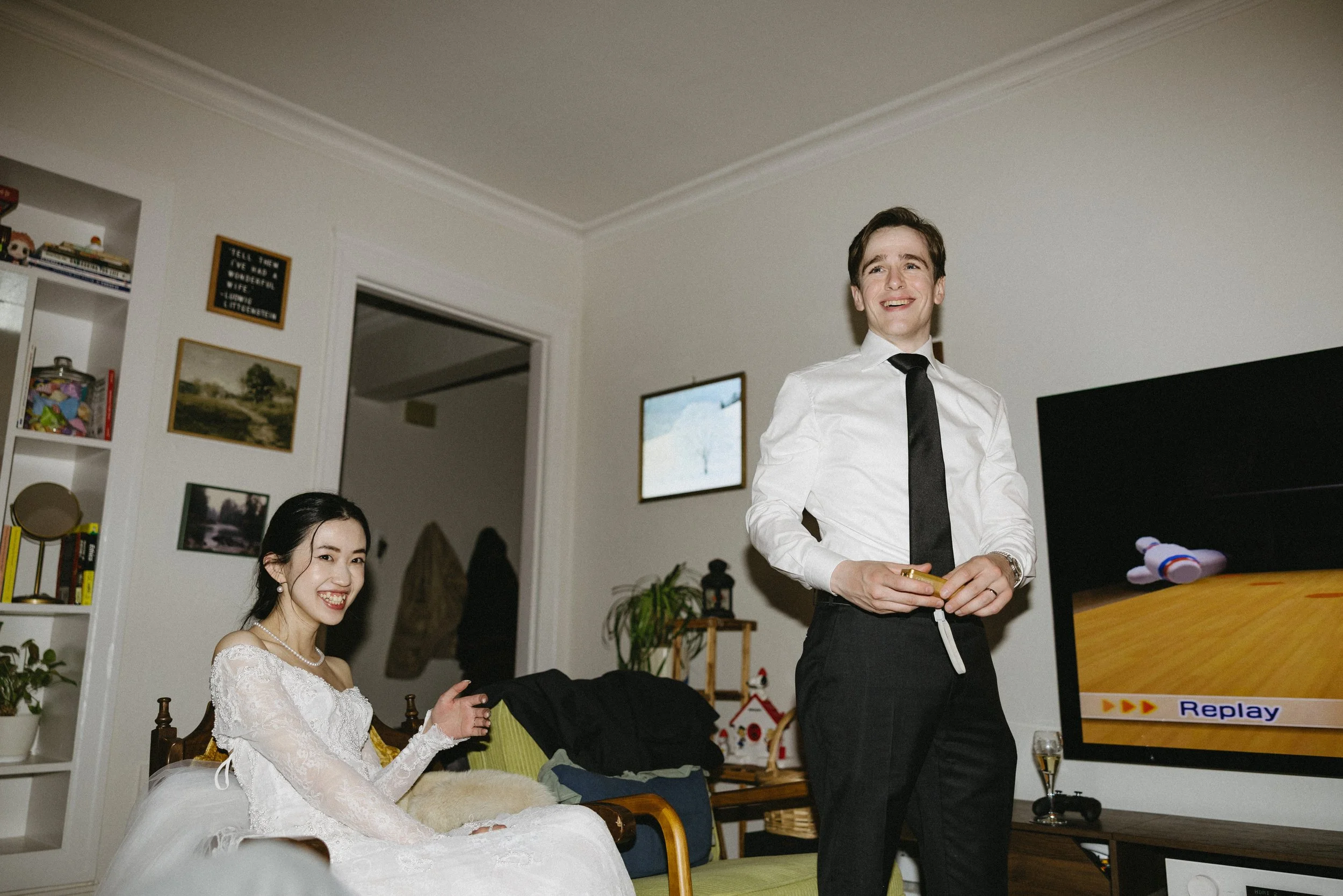 A young woman in a white dress sitting on a green couch smiling, next to a man in a white shirt and black tie standing and holding a game controller in a living room with a TV, bookshelf, and wall art