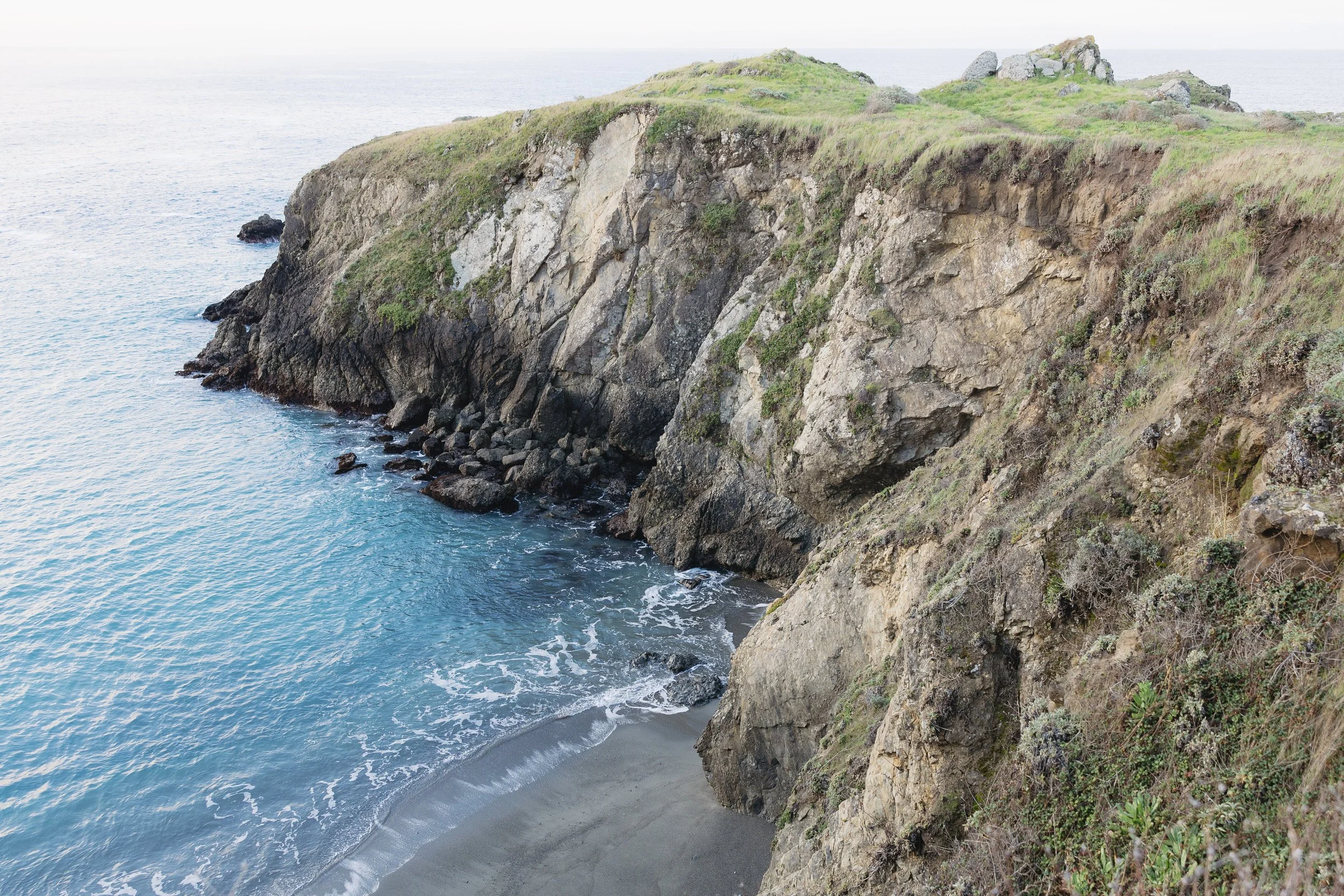 Cliffside overlooking a beach with waves crashing against rocks and sand, grassy top of the cliff, ocean in the distance. wedding venue with dramatic coastline views.