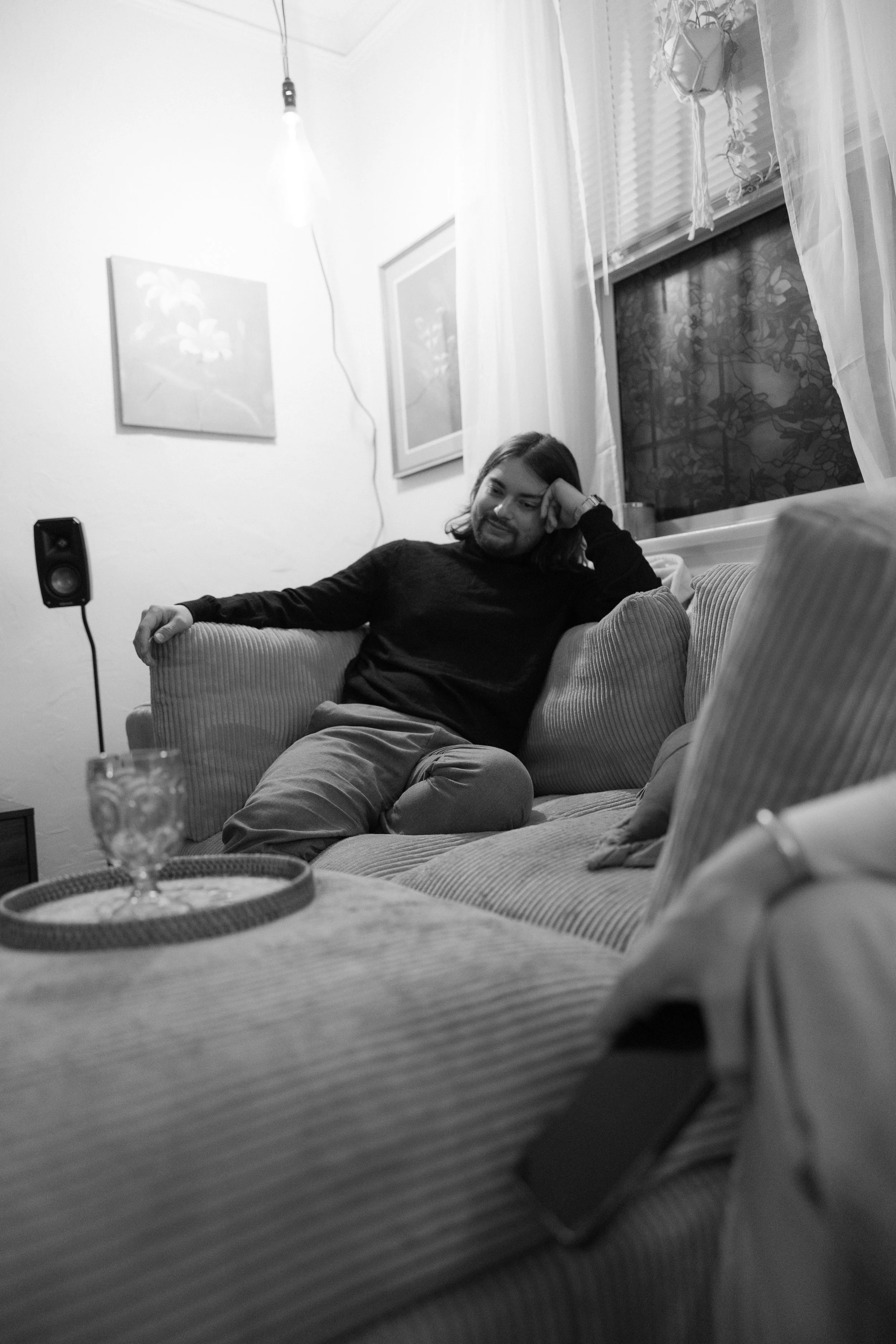 A man with shoulder-length hair and a beard, wearing a black shirt and light-colored pants, sitting on a corduroy couch in a living room, looking relaxed with his head resting on his hand. There is a window with blinds and curtains behind him, and fr