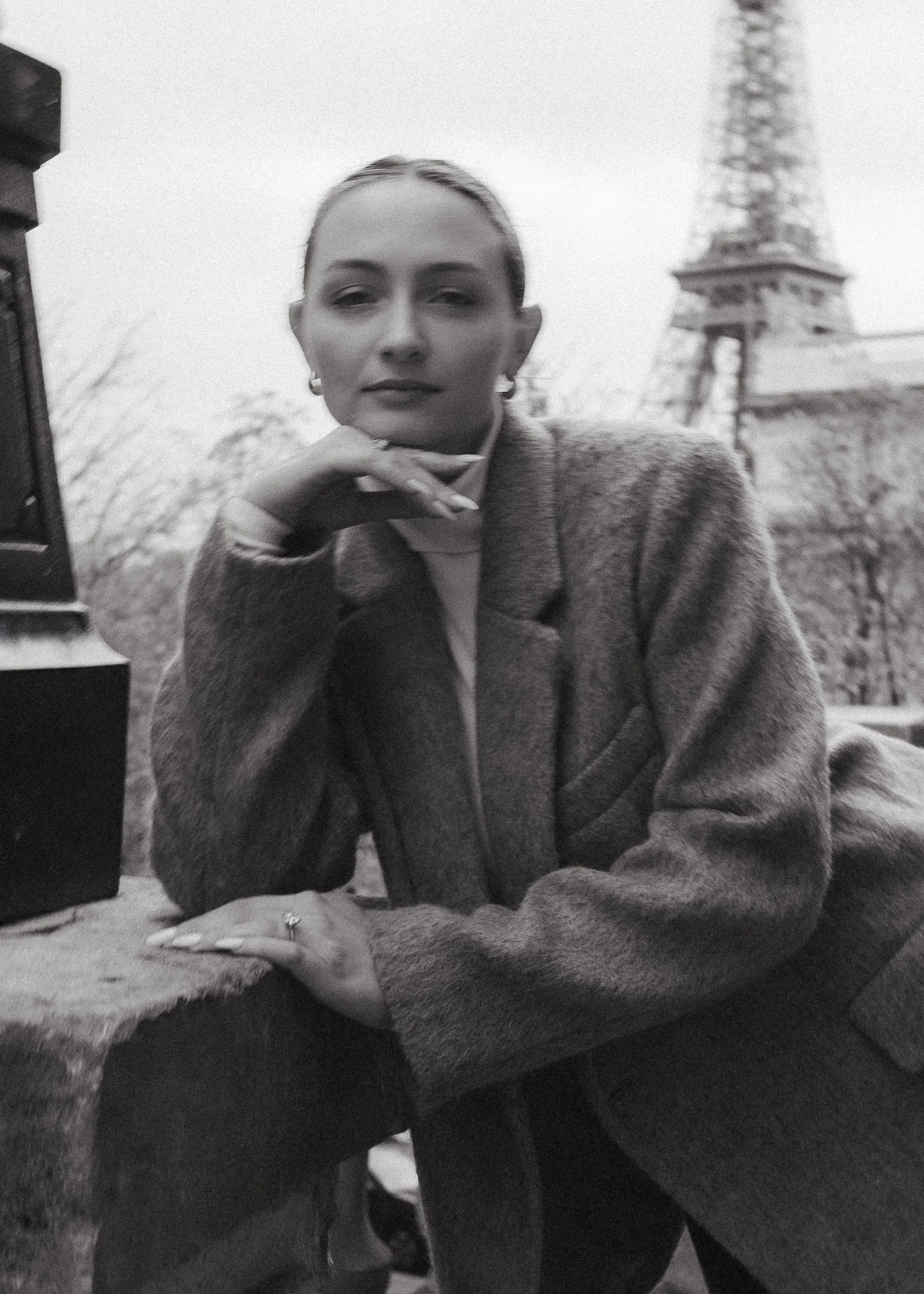 Black and white photo of a woman sitting outdoors with the Eiffel Tower in the background. She is resting her chin on her hand, looking at the camera, wearing a coat and earrings.